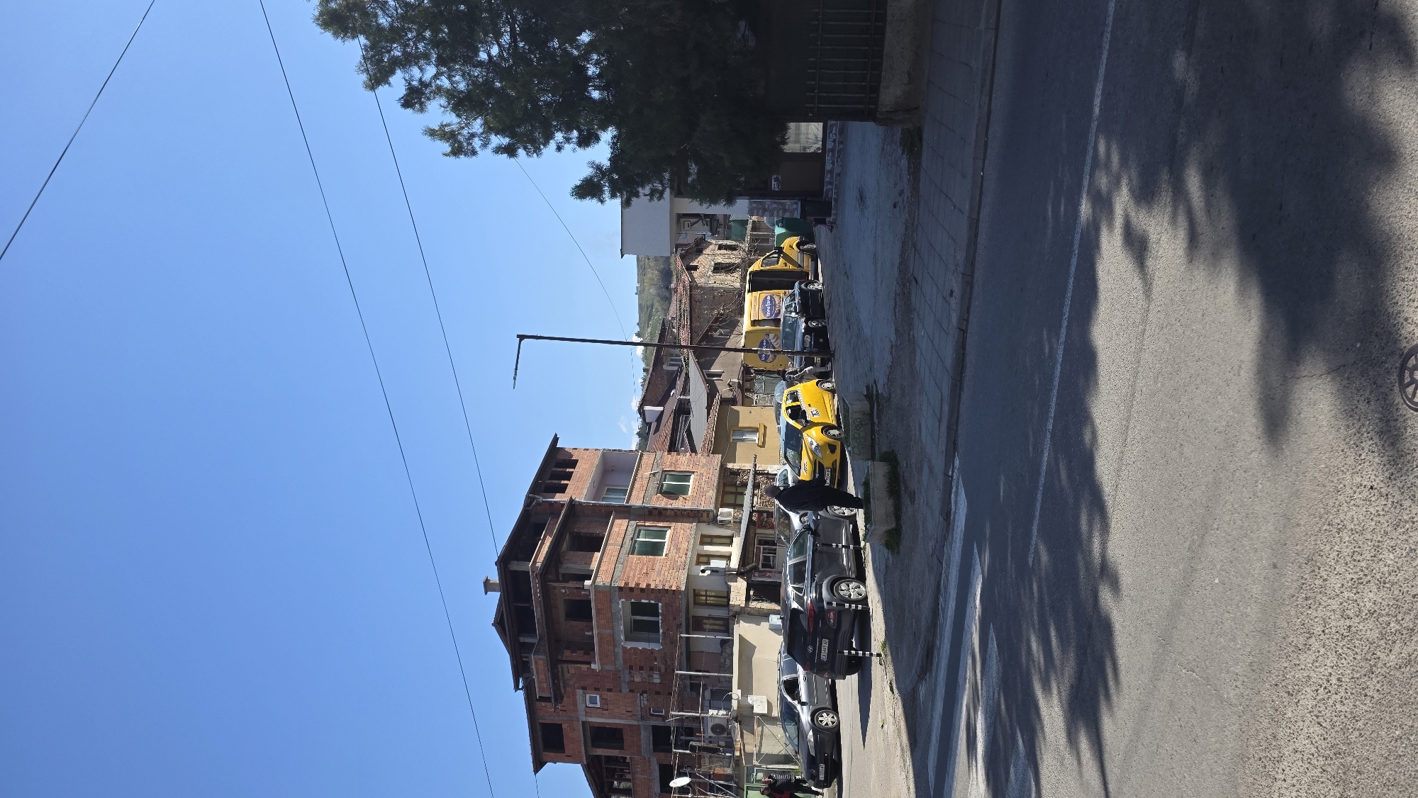 The image shows a street scene in Blagoevgrad, Bulgaria. A multi-story building under construction with exposed brick is on the left side of the street. Parked cars line the right side of the street. A yellow taxi is among the parked vehicles. A street lamp stands near the building. Trees provide shade, casting shadows on the road. The sky is blue.