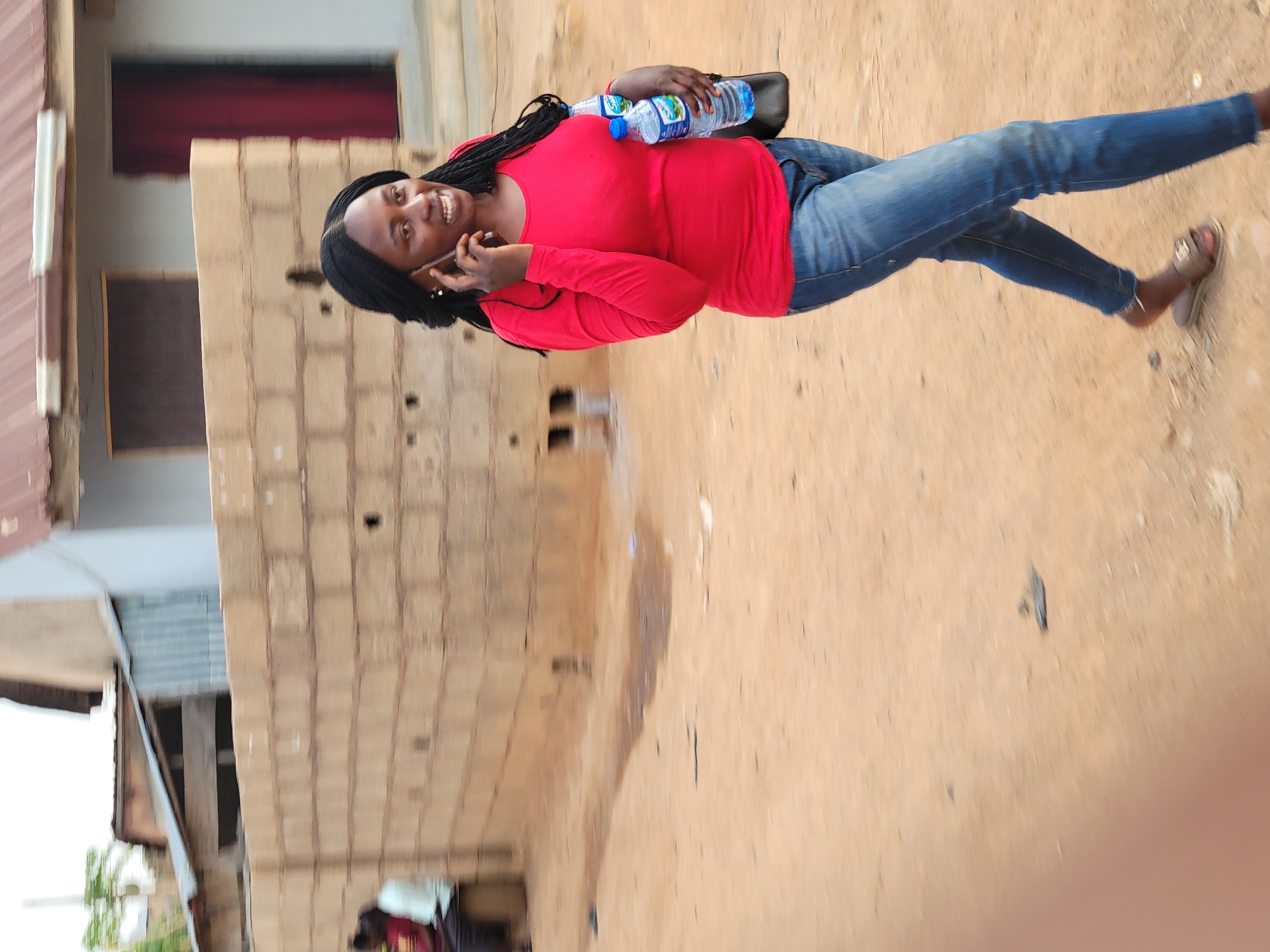 A woman stands on a dirt road in Jos, Nigeria, holding water bottles and a mobile phone to her ear. She wears a red long-sleeved shirt, blue jeans, and sandals. Behind her is a partially visible building constructed of cinder blocks and corrugated metal. The ground is a light brown color.