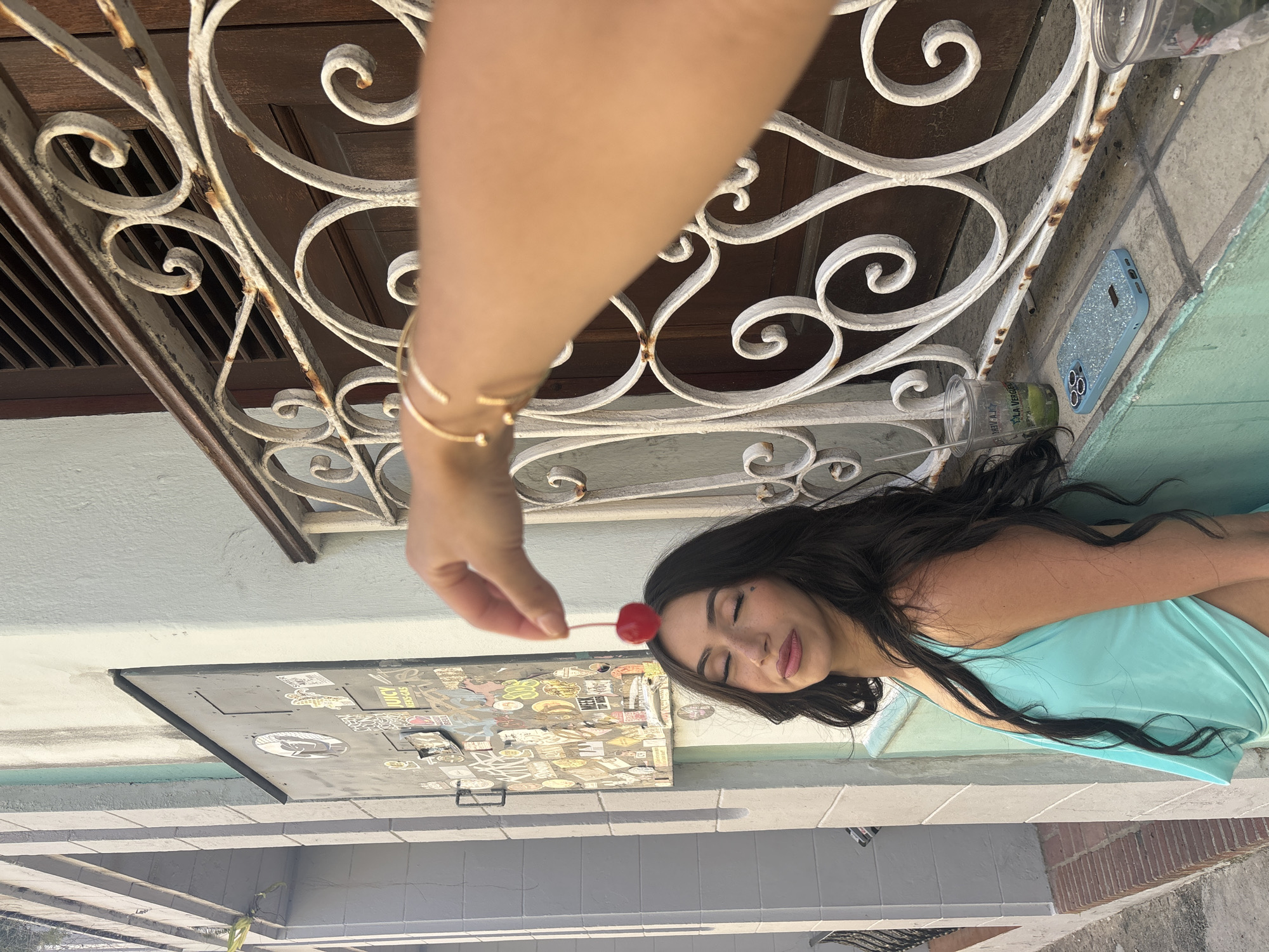 A young woman with long dark hair and closed eyes stands with her head tilted up, a cherry balanced precariously on her forehead. She wears a turquoise tank top with a low neckline. Her eyes are adorned with a small heart-shaped sticker or tattoo under her left eye. Beside her, a metal grate covers a wooden door. A hand with a gold bracelet is visible in the foreground, holding the cherry. On a ledge below the grate, a blue glitter-covered phone case sits next to two plastic cups containing a green liquid and ice. The background shows a textured, pale wall with a dark metal panel covered in stickers, suggesting an urban environment. The architectural style and the presence of graffiti-style stickers on the panel are consistent with the character of Old San Juan.