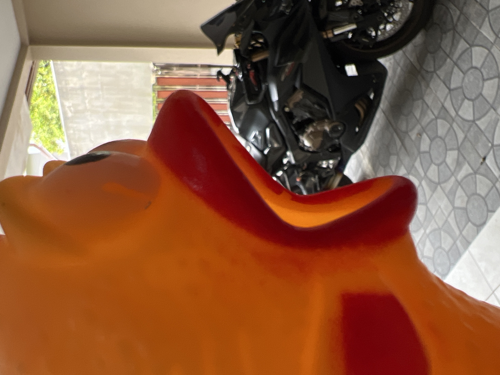 A close-up, low-angle shot shows a large, bright orange cartoon duck toy in the foreground, with its mouth wide open and a red beak. Behind the duck, a black sport motorcycle is parked on a tiled floor. The motorcycle is angled towards the right side of the frame. The background features a concrete wall, some greenery, and a ceiling or overhang. The scene appears to be outdoors, possibly in a covered parking area or a patio, consistent with a residential setting in Choeng Thale, Thailand. The lighting suggests it is daytime.