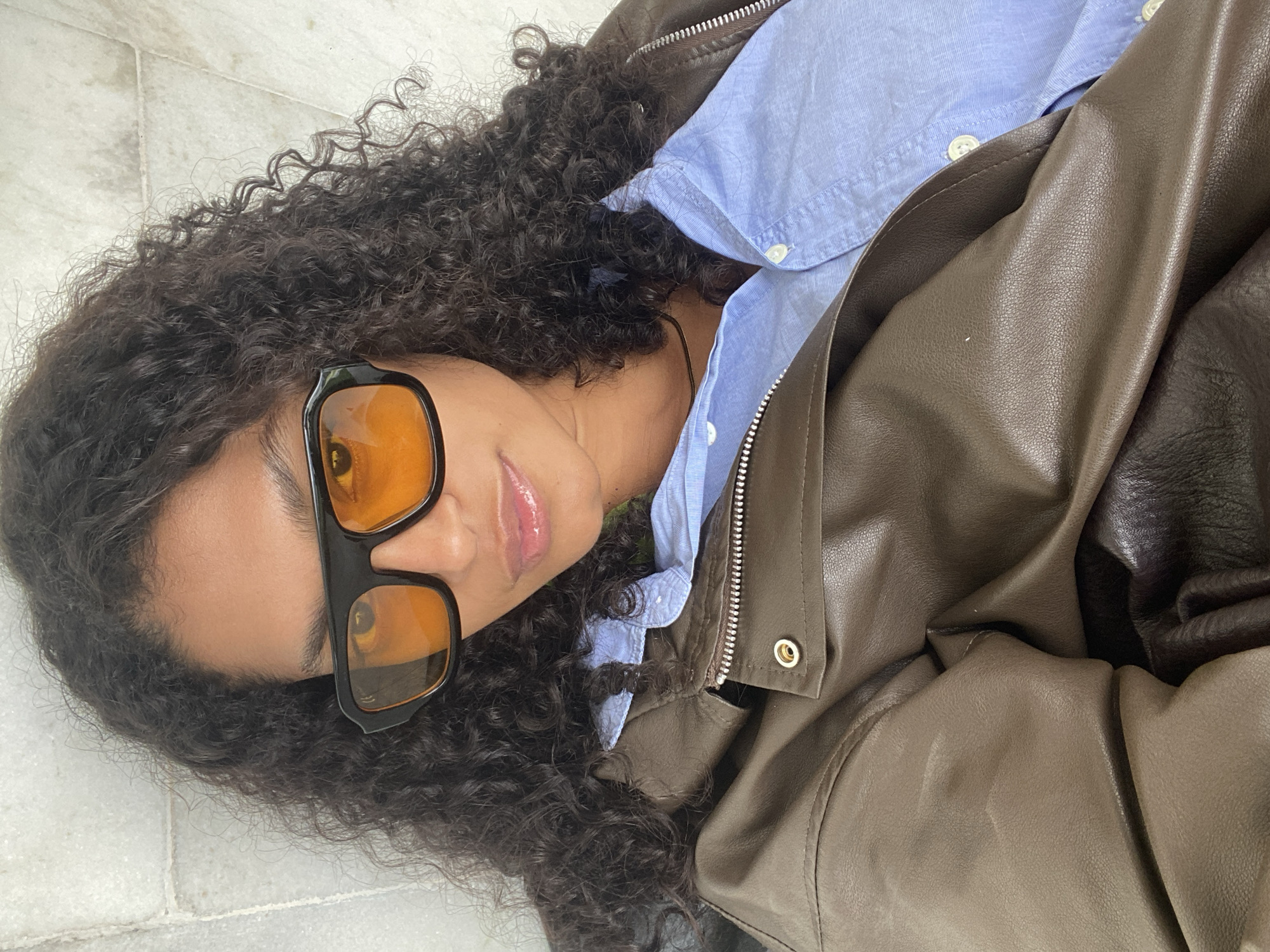 A woman with curly dark hair, wearing black sunglasses with orange lenses, a light blue collared shirt, and a brown leather jacket, lies on a tiled floor. The location is not identifiable as being in São Paulo, Brazil, from the provided image.