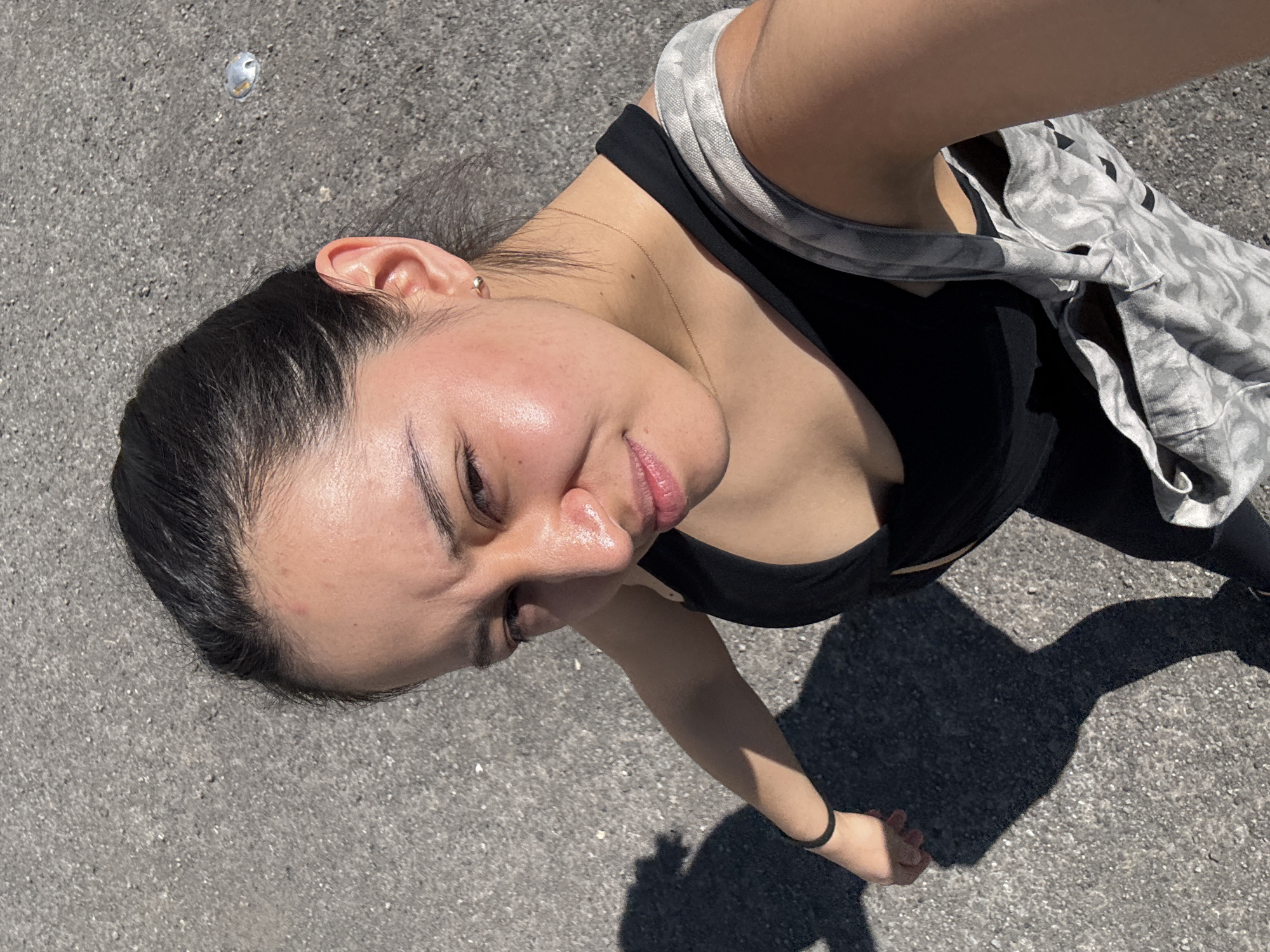 A high-angle outdoor photograph captures a young person from an overhead perspective, looking upwards toward the camera. The subject, who appears to be female, is positioned on grey asphalt or concrete pavement, which shows small aggregate details. Her head is tilted, and she has dark hair pulled back, revealing a small gold stud earring in her left ear. Her facial features include strong eyebrows, slightly flushed and shiny skin (possibly from sun exposure or exertion), and full lips with a soft, relaxed expression.
She is wearing a black sports top or bra, and a light-colored, possibly tie-dye or patterned, shirt or jacket is casually draped over her shoulders. A thin gold chain necklace is visible around her neck. Her right arm is visible, bent at the elbow, with a black hair tie on her wrist.
The scene is brightly lit by direct sunlight, casting sharp, distinct shadows of the person and her arm onto the ground, indicating it is likely midday or early afternoon on a clear, sunny day. A small, shiny, metallic object (possibly a piece of foil or a small pebble) is visible on the pavement near the top left of the frame. The overall setting suggests an urban or suburban outdoor environment, specifically in San Bernardino Tlaxcalancingo, Mexico. There is no visible text in the image.