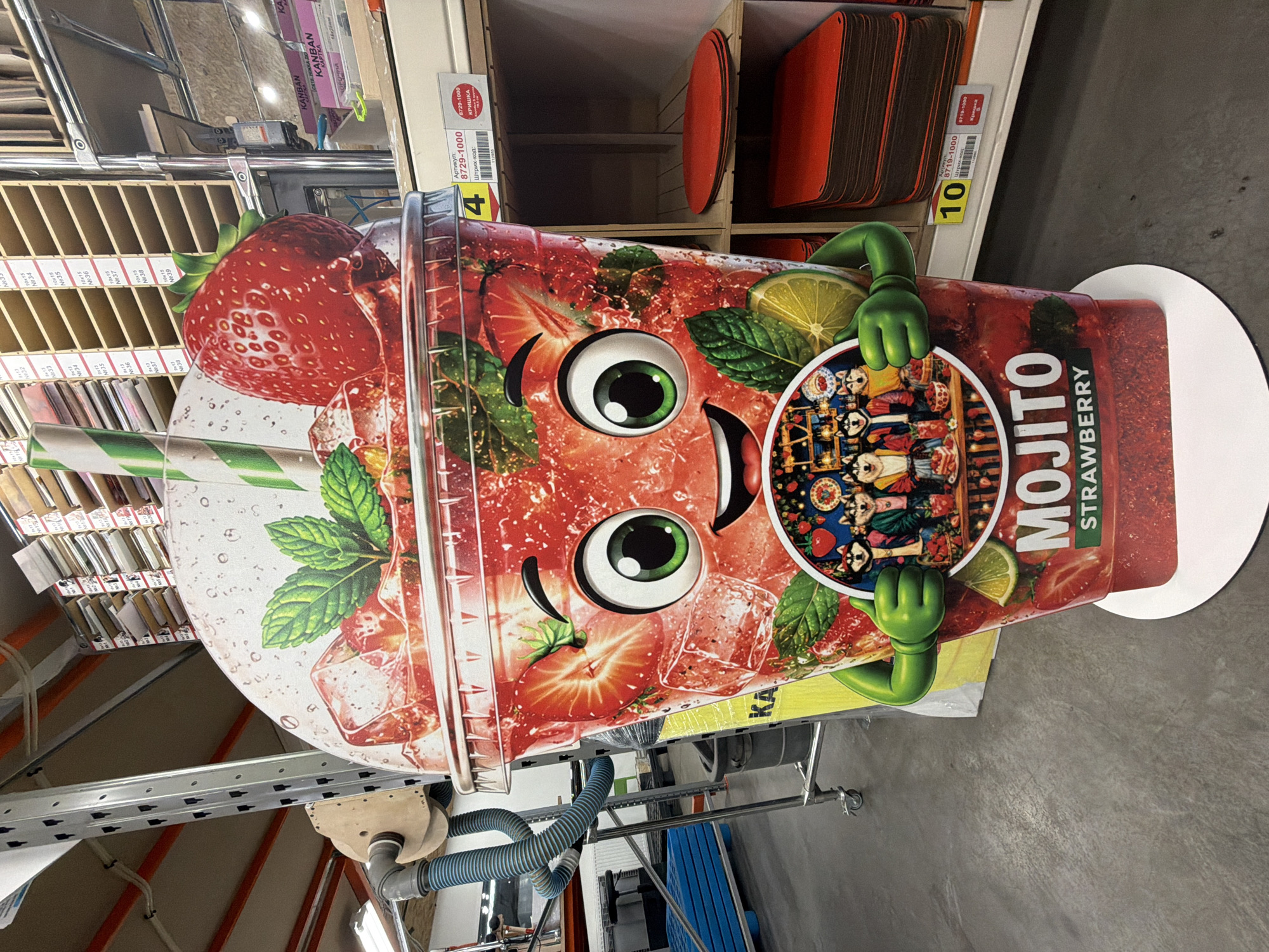 A vibrant, full-sized cardboard standee, shaped like a strawberry mojito drink, dominates the foreground of an indoor, commercial setting. The standee features a cartoon-like drink with a large, smiling face, big green eyes, and green gloved hands holding the sides of the "cup." The drink design includes prominent strawberries, mint leaves, lime slices, and ice cubes. A circular emblem on the standee displays small, colorful cartoon dog-like figures, possibly dressed in traditional Mexican attire or associated with Día de los Muertos, against a detailed background. The words "MOJITO STRAWBERRY" are clearly visible on the side of the standee.
The standee is positioned in what appears to be a retail store or warehouse in Ternopil, Ukraine. In the background, metal shelving units are visible, stocked with various products. One section of shelves holds numerous small, rectangular items, possibly craft supplies or art components, with labels such as "10x15" and sequentially numbered "N№34" through "N№39," and "N№135." Another shelf area displays stacks of red/brown rectangular boards or panels, with labels indicating "KANBAN" and "КАРТКА" (card). Other visible text on labels includes "Артикул: 8729-1000," "Штрих-код: 8729-1000," and "КРИШКА" (lid/cover), alongside yellow tags with numbers "4" and "10." The floor is concrete, and industrial-style pipes and hoses are partially visible in the lower part of the image, suggesting the functionality of the space. The lighting is artificial, indicative of an indoor environment.
