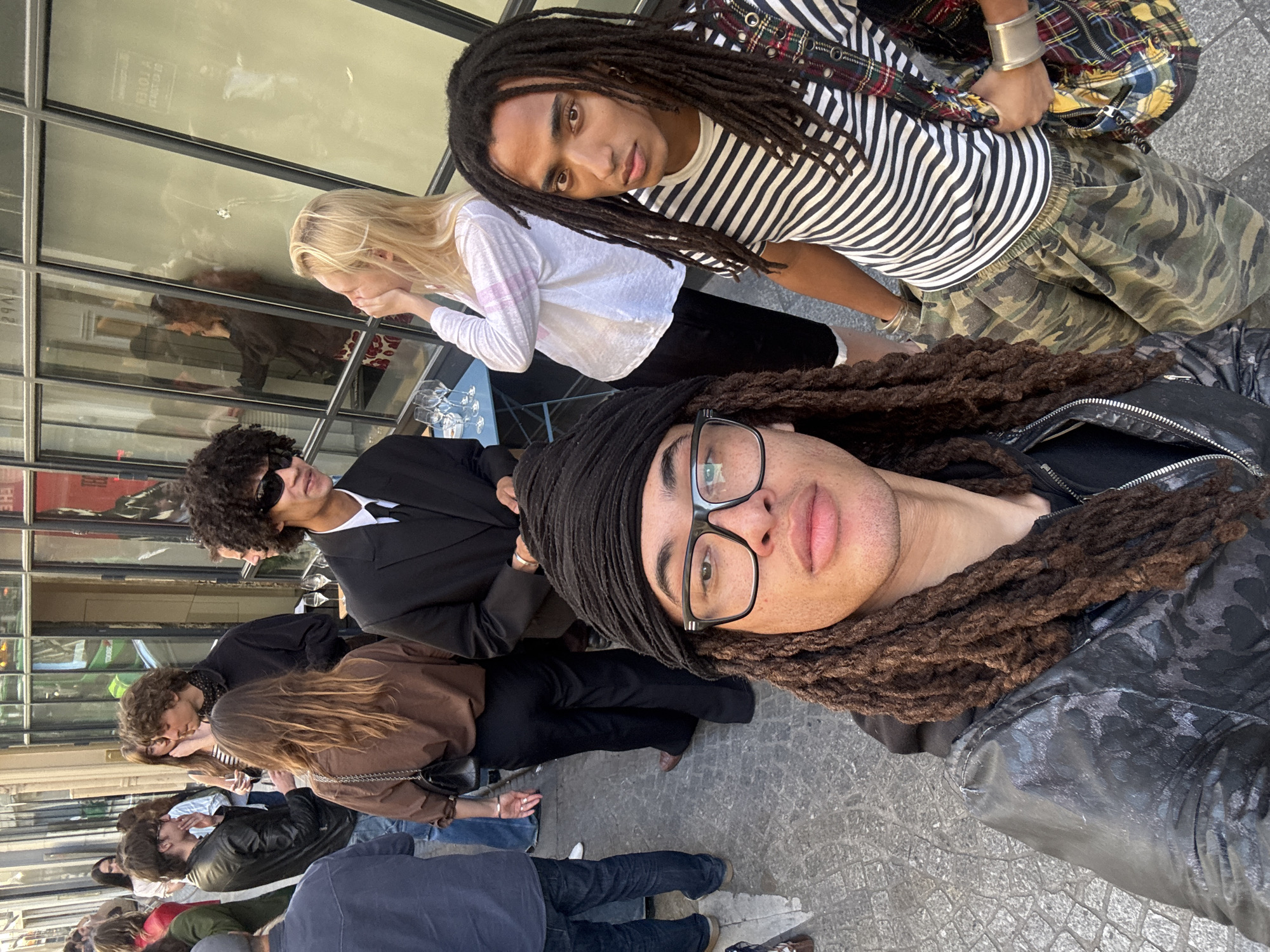 The image shows a group of people, likely in an outdoor setting, with architectural details suggesting they may be in Paris, France.
People: There are several individuals in the scene. Two people are prominently featured in the foreground, possibly taking a selfie or being the main subjects of the photo. One of them is wearing glasses. Additional individuals are seen in the background, standing or sitting. There is a woman with blonde hair and others with dark hair, with varying hairstyles. They appear to be dressed in contemporary clothing.
Objects: The environment includes an exterior facade with a glass front, possibly a store or restaurant, reflecting the surrounding environment and some of the individuals in the scene. There are tables with glasses, suggesting an outdoor dining or café area.
Actions: The foreground subjects appear to be posing for the photograph. People in the background may be dining, conversing, or waiting in line.
Environment: The scene appears to be an outdoor area in a city, with potential clues suggesting it may be in Paris, France, based on architectural styles and the general ambiance. The flooring appears to be cobblestone or paving stones.
Interactions: The primary interaction is between the individuals in the foreground, possibly taking a photo together. Other interactions may involve conversations, dining, or observing the surroundings.