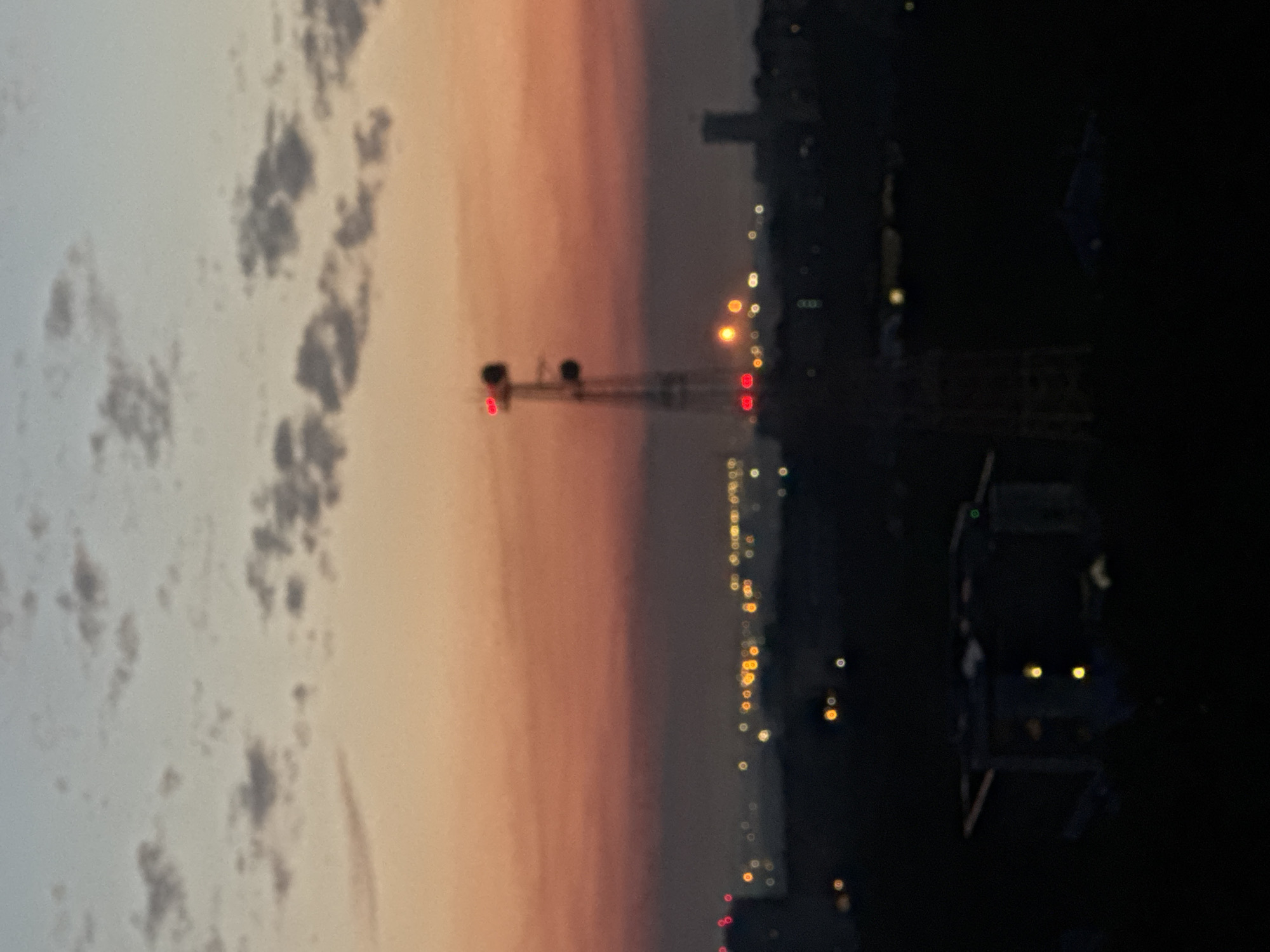 This is a slightly blurred, high-angle, long-shot photograph of a city skyline at dusk. The sky is a gradient of pale blue at the top, transitioning to a soft orange and pink hue near the horizon, with scattered, wispy clouds. A prominent telecommunications tower, lit with red lights at its top and mid-section, stands in the middle ground. Below the tower, the city lights are visible as blurred yellow and white orbs, suggesting a dense urban area. Silhouetted buildings and structures form the dark foreground. The overall scene depicts an urban environment at twilight, possibly in the city of Ufa, Russia, given the context. No people or specific actions are discernible due to the distance and blur.