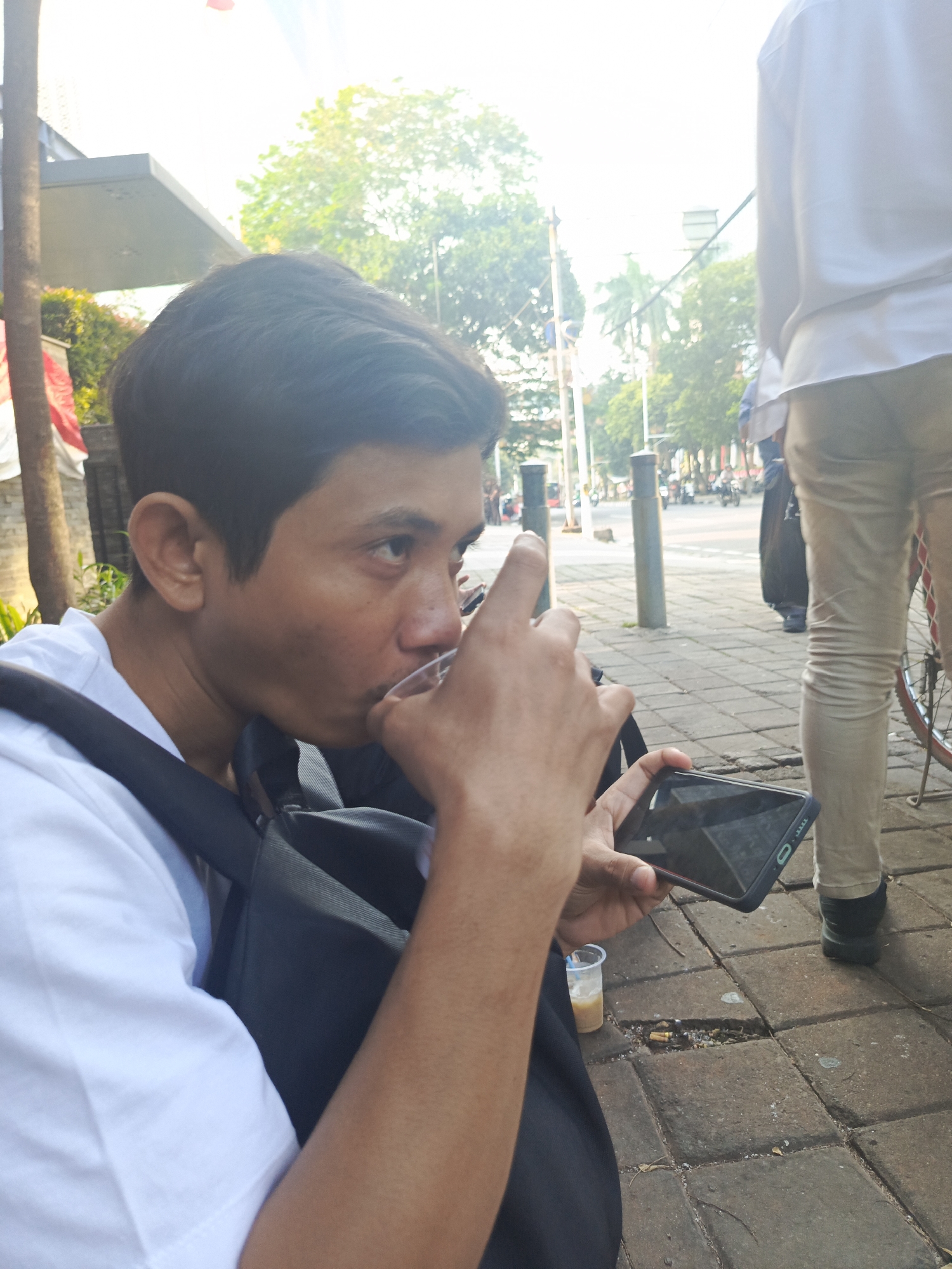 A young man is sitting on a tiled sidewalk in Central Jakarta, Indonesia. He is wearing a white t-shirt and has a black backpack on. He is holding a clear plastic cup to his lips, drinking from it, and looking to his right. In his left hand, he holds a black smartphone. A partially filled plastic cup with a light brown liquid is on the sidewalk near him. The background shows trees, buildings, and traffic poles, indicating an urban environment. Another person, wearing a white shirt and light-colored pants, is standing to the right, with a bicycle wheel visible next to them. The man's left hand is raised, with his index finger extended. The scene appears to be daytime, with bright sunlight filtering through the trees.