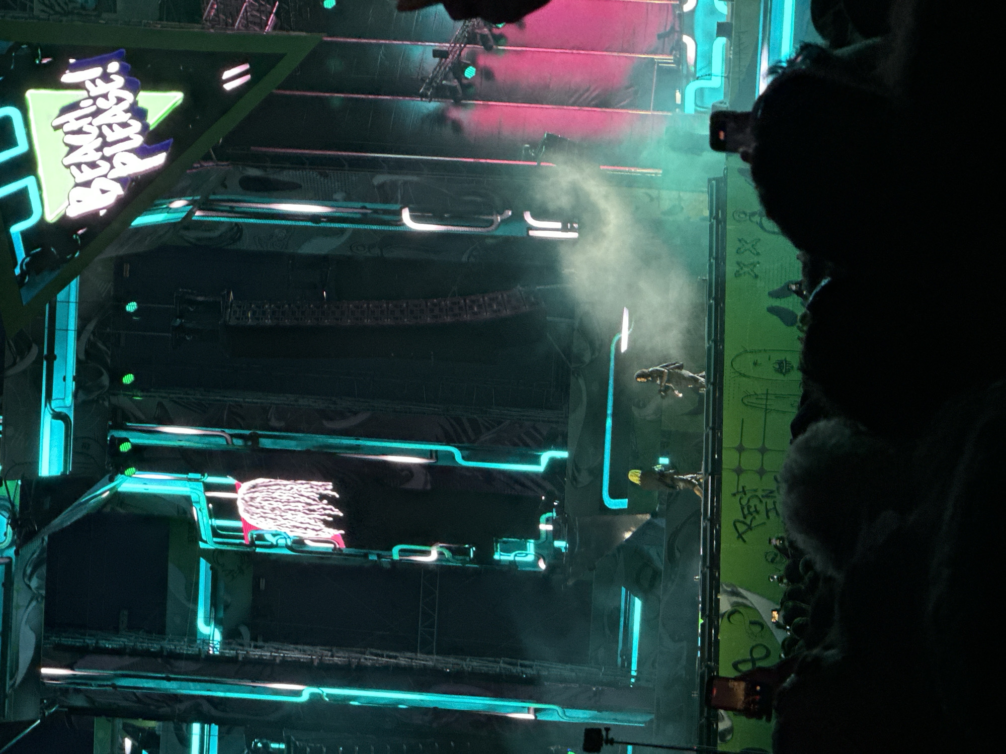 This image captures a night scene at the Beach Please festival in Costinești, Romania. The stage is lit with vibrant neon blue and pink lights, creating a futuristic and energetic atmosphere. A large, triangular sign with "BEACH PLEASE!" emblazoned in white and purple lettering is prominently displayed. On the stage, two figures are visible: one with bright yellow hair, wearing a dark outfit, and another in darker attire. Smoke or fog machines are actively producing a cloud of mist that swirls around the performers. In the foreground, a crowd of people are silhouetted against the stage lights, with some holding up their phones to capture the event. The overall impression is that of a lively music festival in full swing.