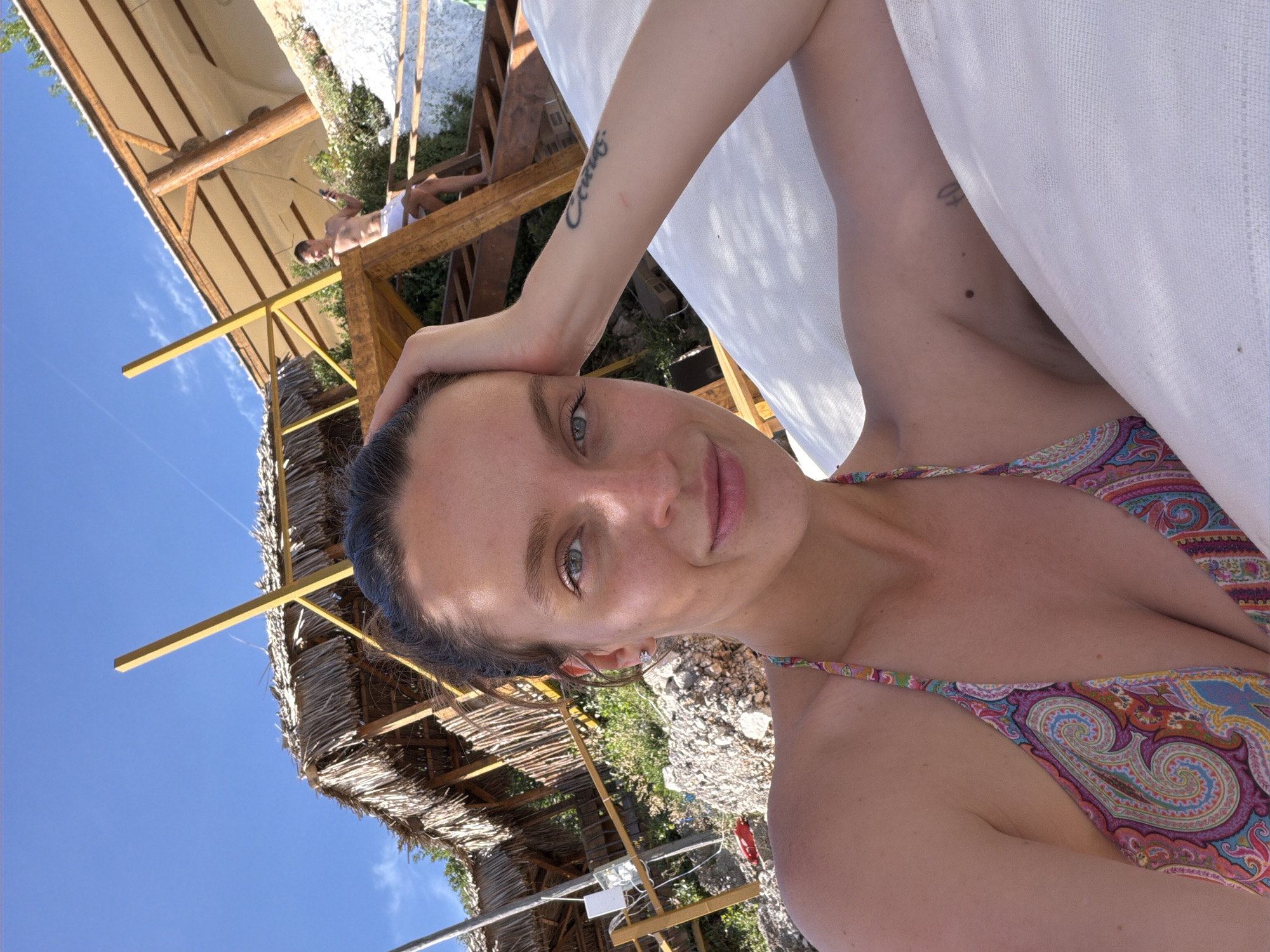 The image shows a woman taking a selfie. She is wearing a patterned bikini top and has her hand on her head. Her skin is fair, and she has a tattoo on her arm. In the background, there is a wooden structure with a thatched roof and a person in the distance wearing white shorts. The sky is blue. The location is likely near a beach with sunshades in Budva, Montenegro, due to the thatched roof structure and the general appearance of the landscape.