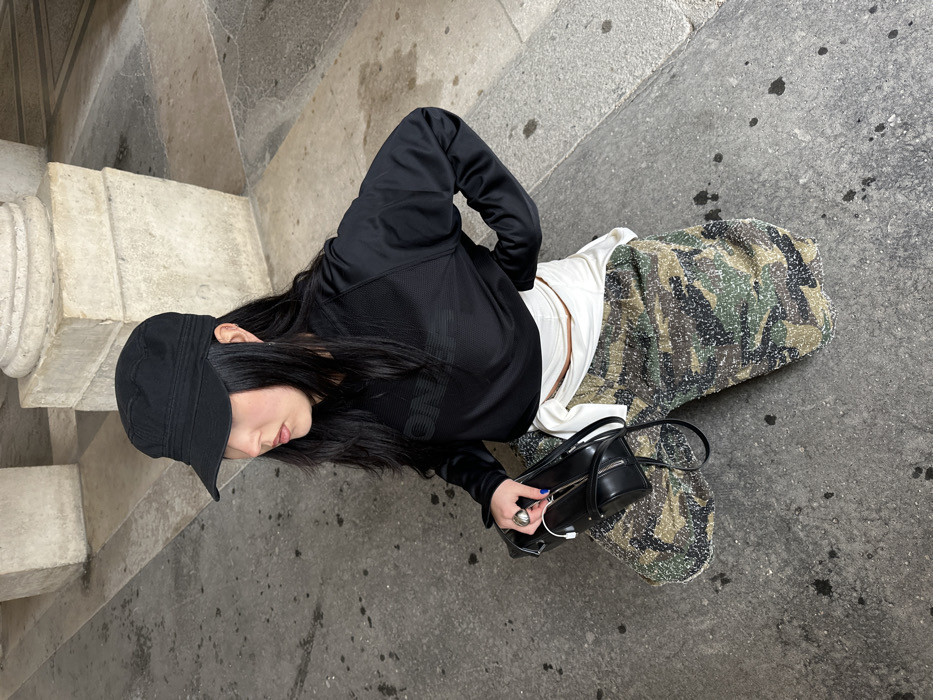 A female individual is depicted in a crouched or kneeling position on a grey paved surface, located within a public arcade or portico in Paris, France, indicated by the partial view of a classical light-colored stone column in the background. She wears a black cadet-style cap, a black long-sleeved mesh top with a subtle "ONLY" graphic across the chest, and textured, camouflage-patterned trousers. A white garment is tied around her waist. Her left hand holds a black rectangular handbag, from which a white wired object, resembling earbuds or a charging cable, protrudes. She wears a silver ring on her left index finger, and her fingernails are painted blue. Her right arm is bent, with her hand positioned on her hip/waist area. She has long, dark hair visible beneath her cap and is looking towards the left side of the frame. The ground exhibits some dark stains.