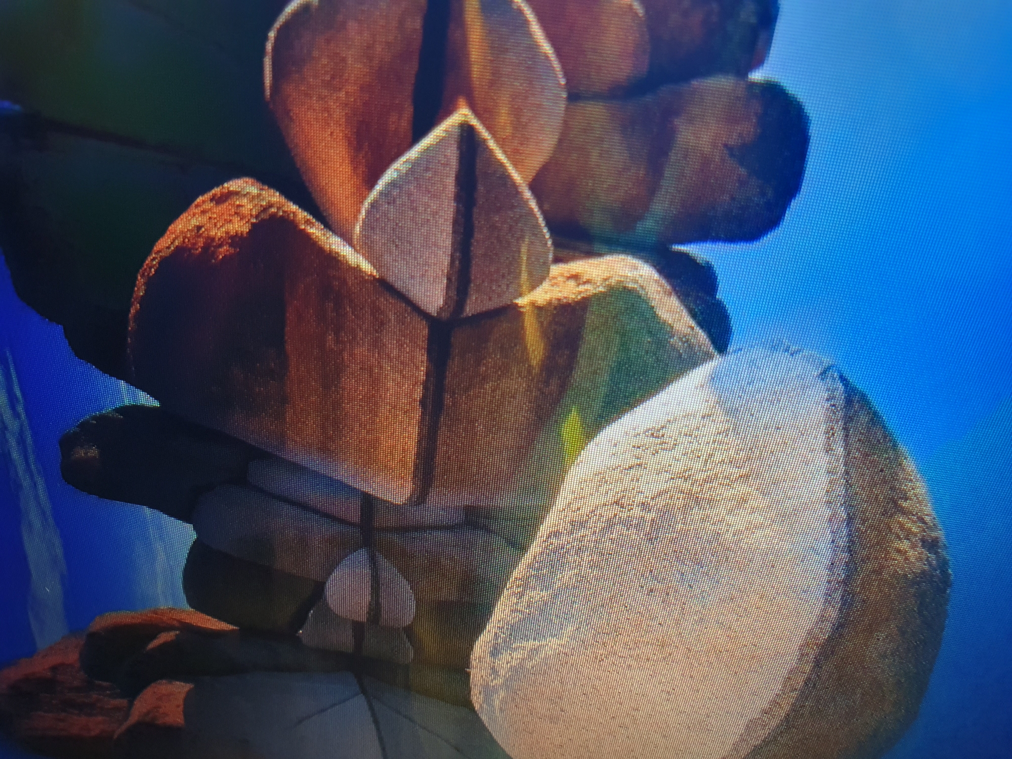 The image displays a close-up, pixelated view of a digital rendering of large, smooth, rounded rocks. The rocks are predominantly earthy tones of brown and tan, with some showing reddish hues. They appear to be clustered together and partially submerged in a reflective, blue surface, suggesting water or a mirrored environment. The lighting creates highlights and shadows on the rock surfaces, emphasizing their texture and form. The background consists of a bright blue sky with subtle horizontal lines, possibly clouds or a stylized sky effect. There are no people or animals visible in the image, and the scene is devoid of any specific identifiable landmarks or environmental features that would place it in Tashkent, Uzbekistan, or any other specific city. The rendering style suggests a digital artwork, game environment, or a similar simulated scene rather than a photographic capture of a real-world location.