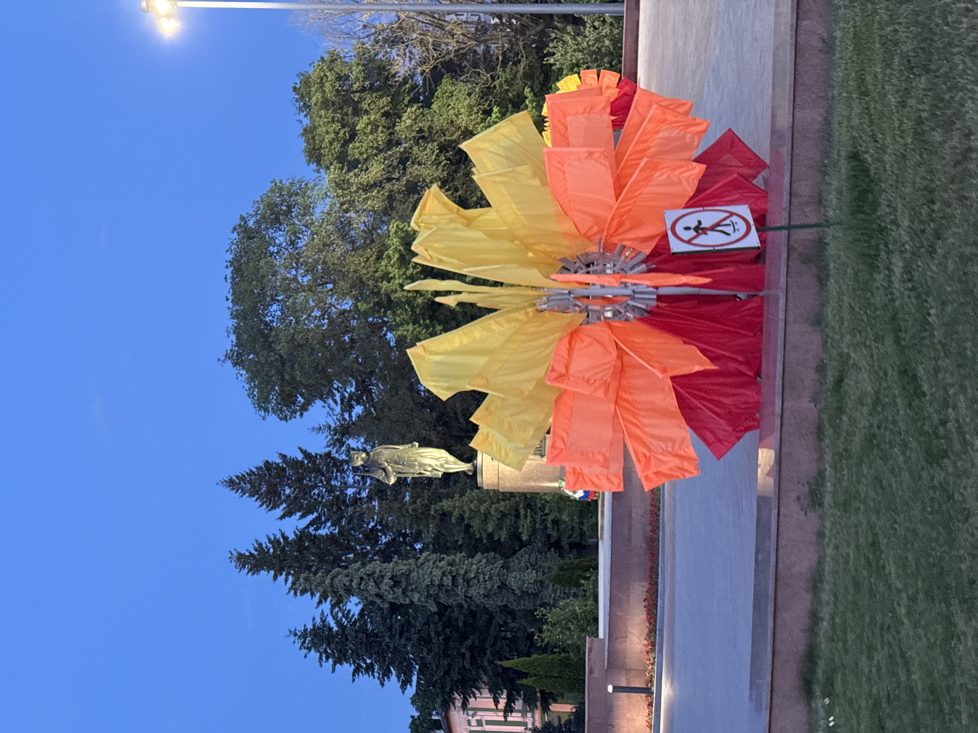 The image captures a public space in Unknown, Unknown, displaying a monument against a twilight sky. Dominating the foreground is a large, decorative flower-like structure composed of red, orange, and yellow fabric petals radiating from a central hub. A sign depicting a crossed-out figure, indicating a prohibited action, is positioned on or near this structure. In the background, a bronze statue of a figure stands atop a stone pedestal, framed by lush green trees, including conifers and deciduous varieties. A street lamp shines brightly in the upper left corner of the frame. The composition suggests a location within a park or a public square, with a manicured lawn extending to the right edge of the image.