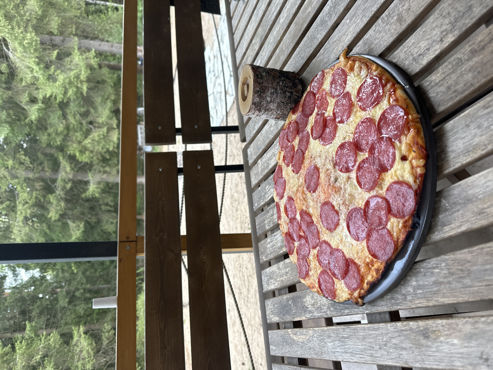 The image shows a pepperoni pizza on a metal tray, resting on a weathered wooden table. Beside the pizza is a cylindrical piece of wood with a hole bored through its center. The table is on an outdoor structure, possibly a porch or deck, judging by the wooden railing and beams visible in the left side of the frame. Beyond the structure, there is a dense forest, which gives the impression that the location might be in a more rural or wooded area. The location is noted as Прикамский, Russia.