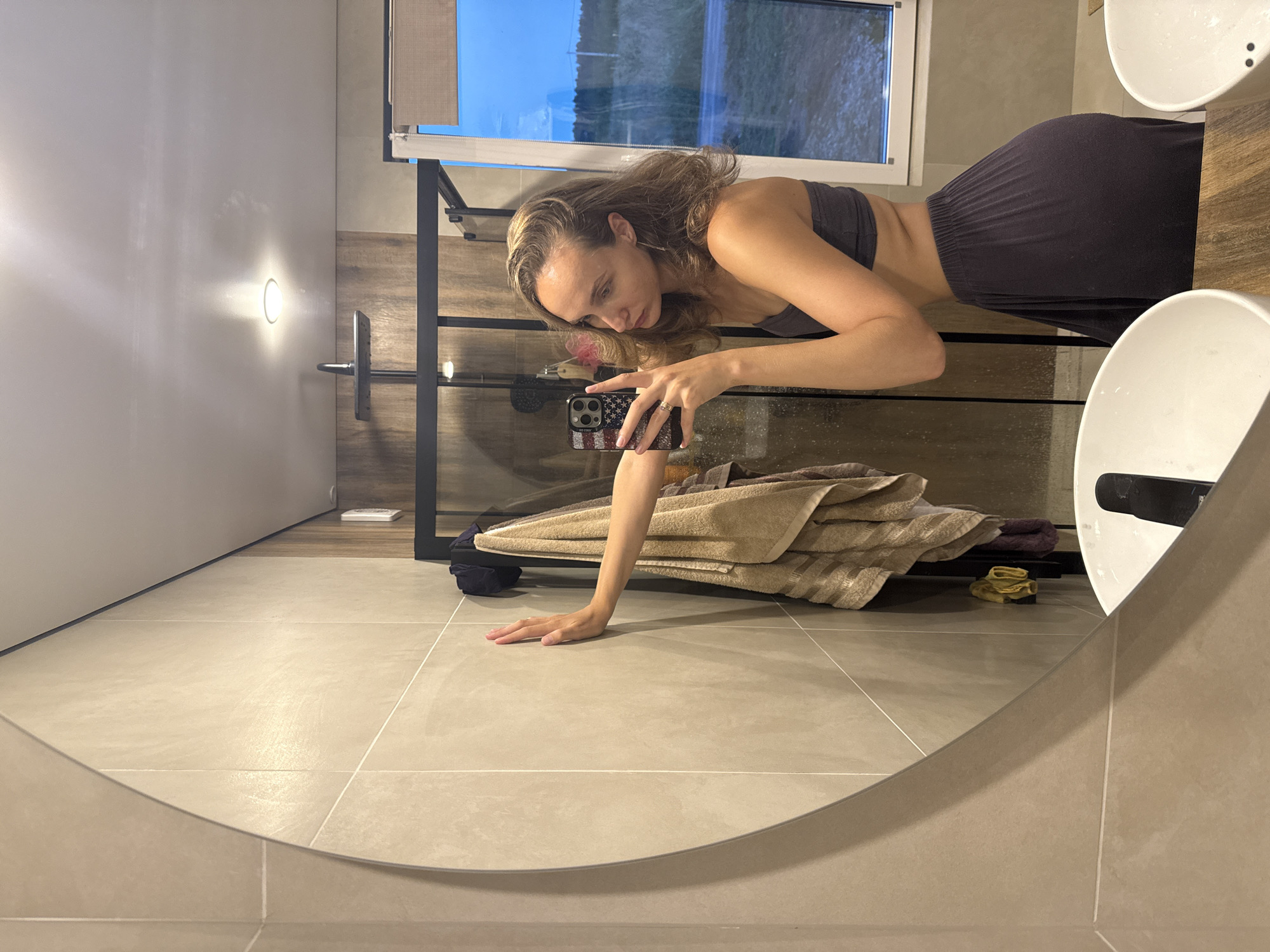 A woman is taking a mirror selfie in a bathroom. She is wearing a gray strapless top and a dark gray maxi skirt. She is leaning against a tiled wall with her left hand. In the mirror, her reflection shows her taking the photo with her phone, which has an American flag case. The bathroom features a large, round mirror, a shower enclosure with a wooden-patterned wall, a sink, and a window with a view of a rural landscape. There are towels hanging on a rack. The lighting is dim, suggesting it might be evening or early morning. The image is captured in the city of Podshivalovo, Russia.