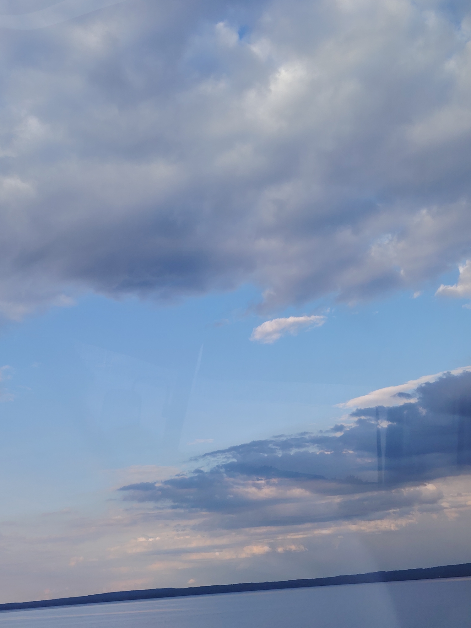 The image shows a vast expanse of sky filled with a variety of clouds, ranging from light wispy formations to heavier, darker masses. The sky transitions from a pale blue at the bottom to a deeper blue towards the top. A body of water, likely a lake or sea, occupies the bottom portion of the image, appearing calm and reflecting the sky. A distant, dark shoreline is visible across the water, suggesting a natural landscape. There are no people or distinct man-made objects in the frame, creating a serene and naturalistic scene. The lighting indicates it is likely late afternoon or early evening. The image is a landscape view, taken from an elevated perspective. The location is identified as Noviy, Russia, consistent with a natural setting that could be found in this region.