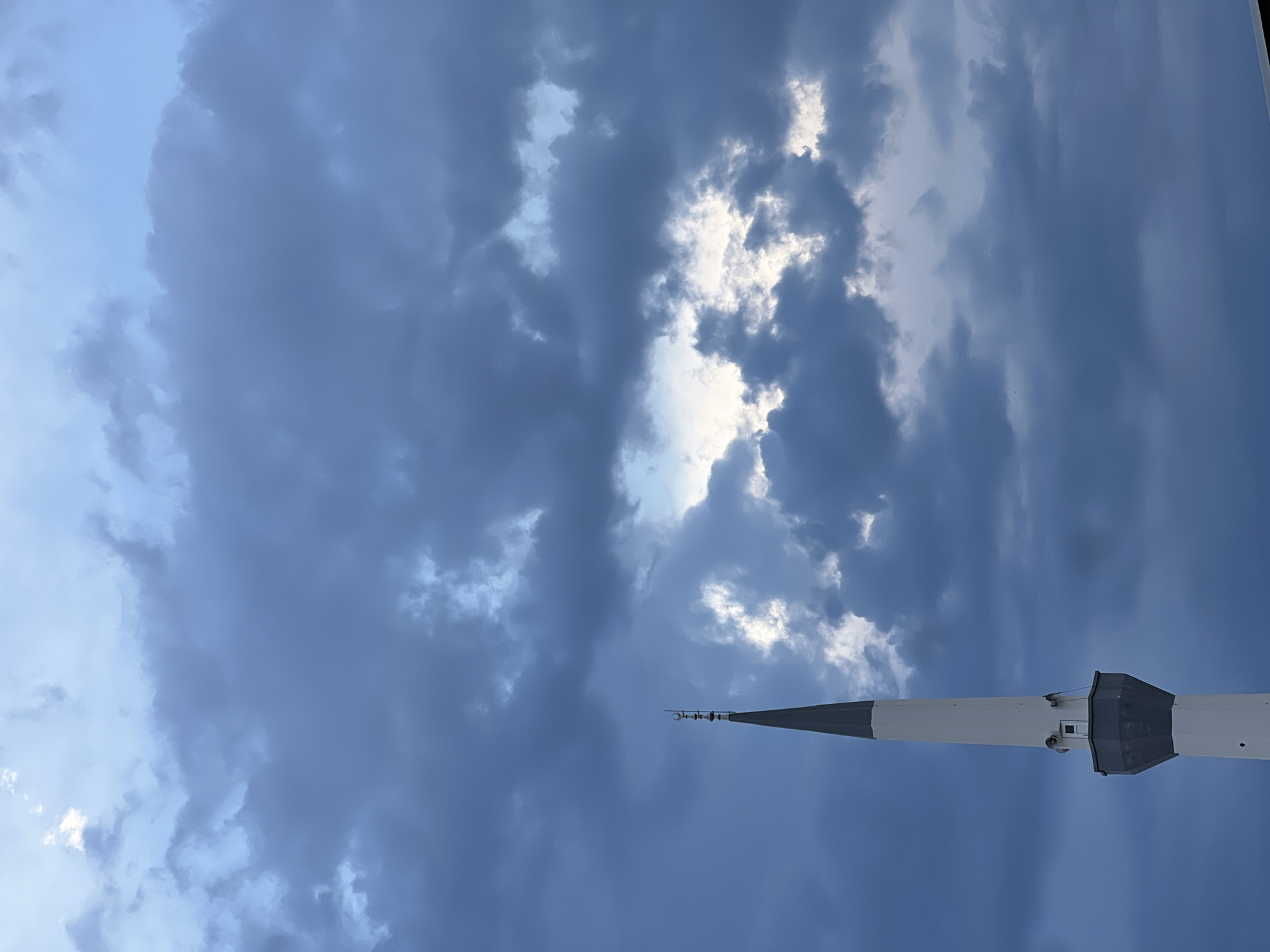 The image shows a minaret from a mosque against a cloudy sky in Antalya, Turkey. The minaret is tall and slender, with a white base that transitions to a gray hexagonal section, and then a pointed white spire topped with a crescent moon. The sky is filled with dark, dramatic clouds with patches of lighter sky visible through them. The overall mood is somber and atmospheric, suggesting perhaps an approaching storm or sunset.