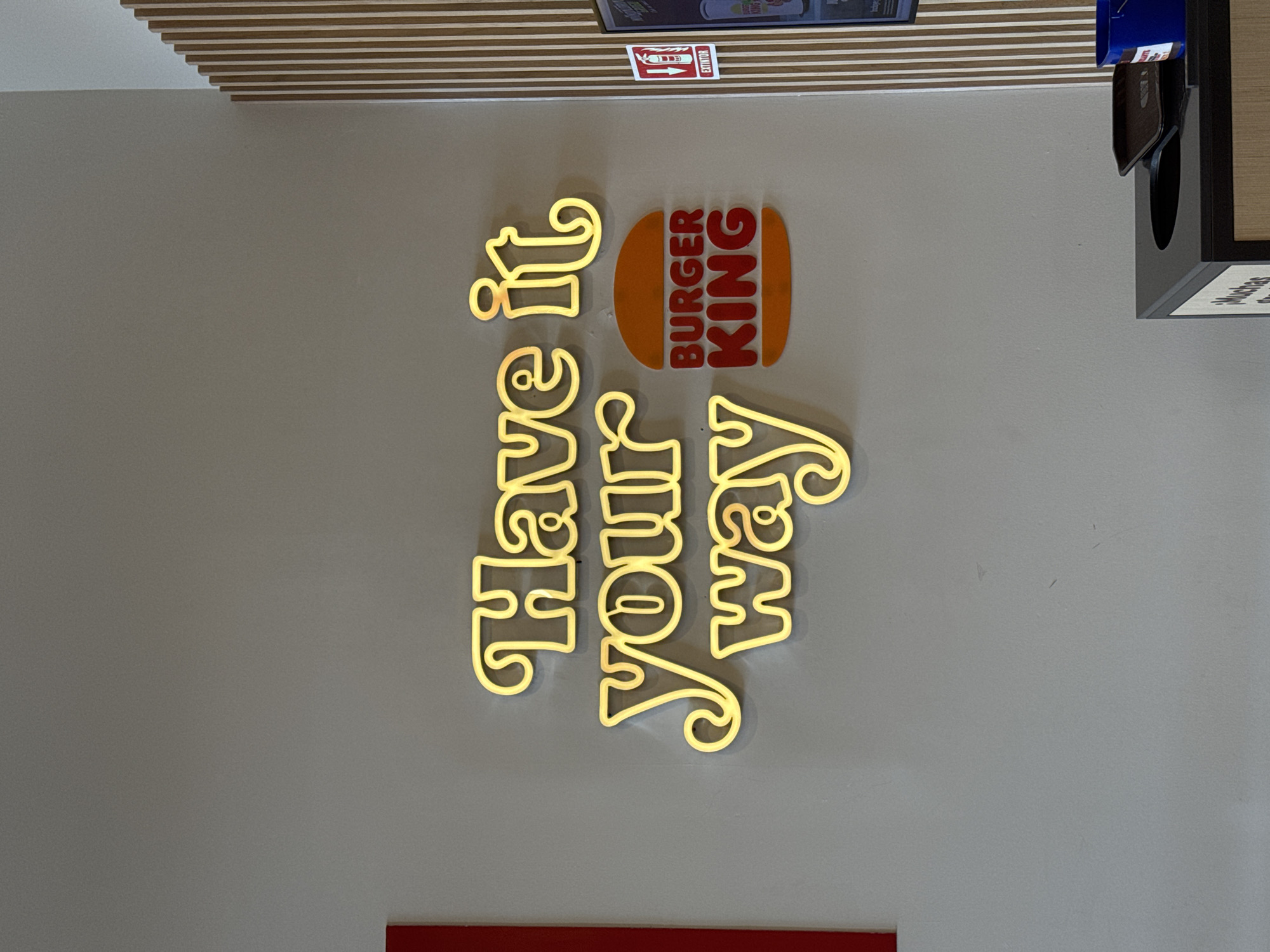 A Burger King logo and slogan ("Have it Your Way") are displayed vertically on a wall. The "Burger King" logo is presented as an orange burger bun with red lettering, and the slogan is in a stylized, illuminated golden font. Above the logo is a small fire extinguisher label, and part of a menu board. To the right of the logo, a trash receptacle with a wooden surface is partially visible. The location is identified as Barrio de Nuevo León, Mexico.