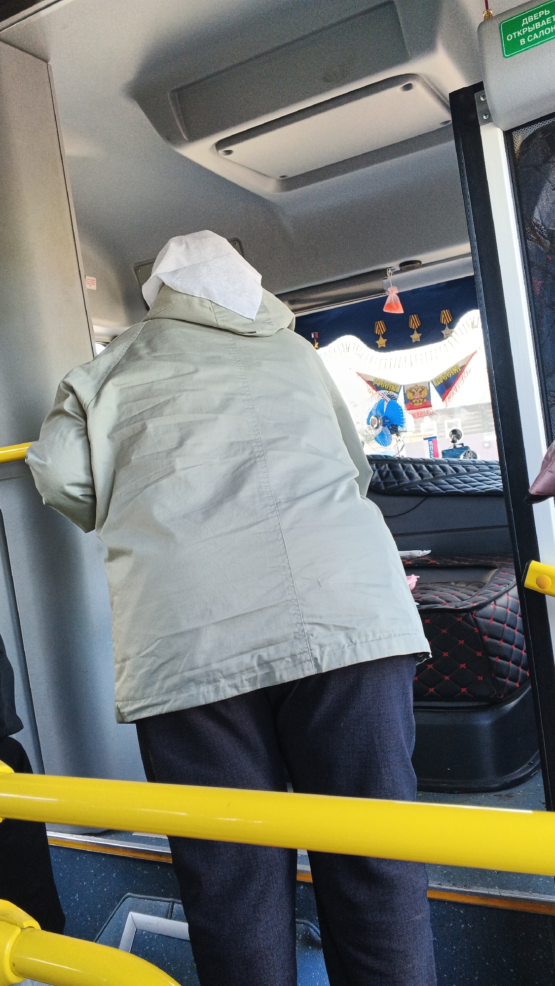 The image shows a rear view of a person wearing a light-colored jacket and dark pants, with a white head covering, standing inside a vehicle, possibly a bus. The person is holding onto a yellow railing. Through the window of the vehicle, Russian flags and other decorations are visible, suggesting a patriotic or commemorative theme. The interior of the vehicle appears to have black and red patterned upholstery on a seat. The location is identified as Киясово, Russia. The scene is illuminated by daylight.