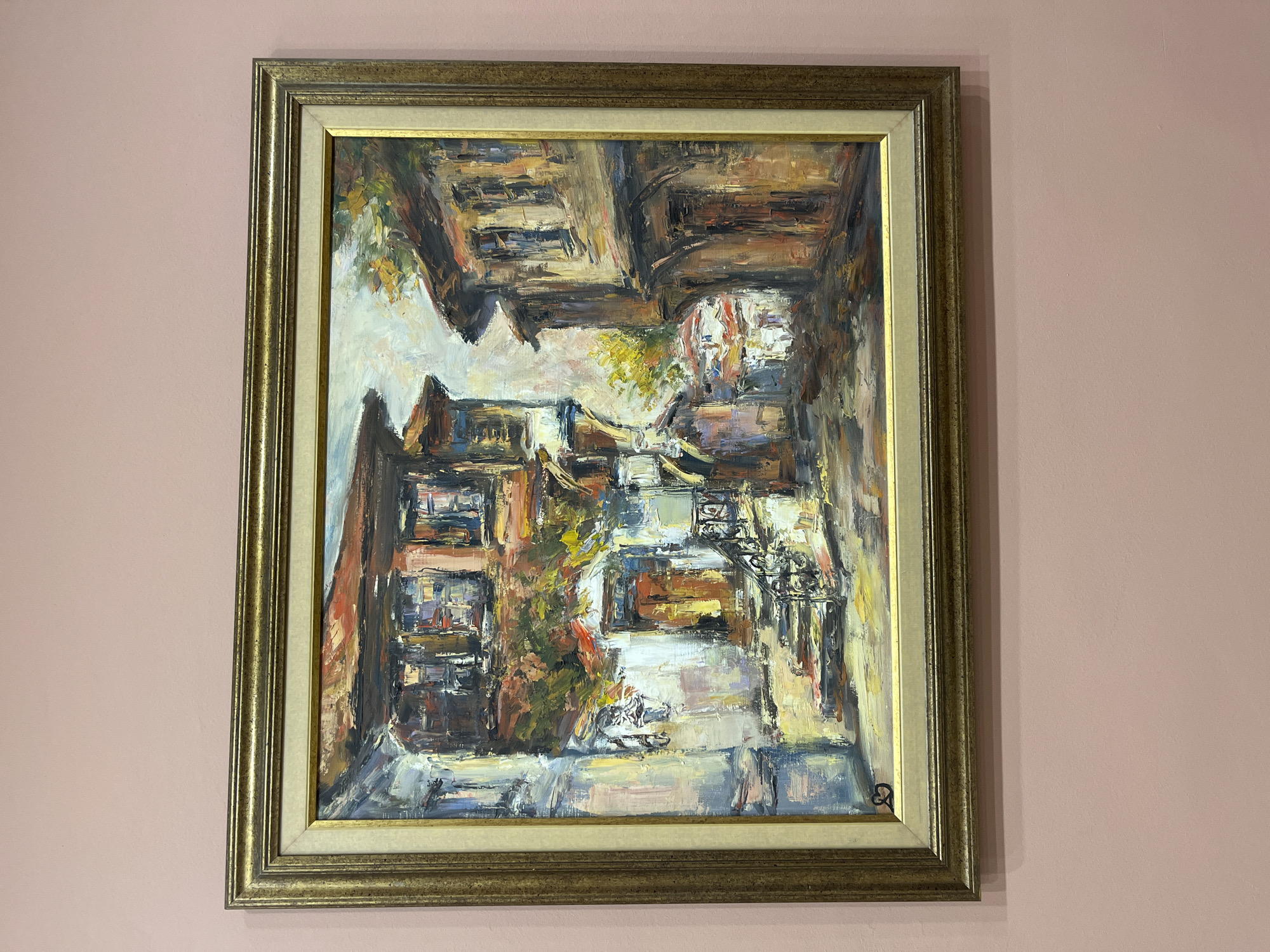 A framed, oil-painted artwork depicting a street scene hangs on a light pink wall. The painting features an abstract interpretation of an urban environment. Buildings are suggested through blocks of color and varying textures. Light and shadow interplay to create depth. The perspective is somewhat skewed, rendering the composition unusual. The scene is possibly located in the city of Plovdiv, Bulgaria. The painting's frame is gold with a lighter inner border. A recognizable signature is visible at the bottom right corner of the painting.