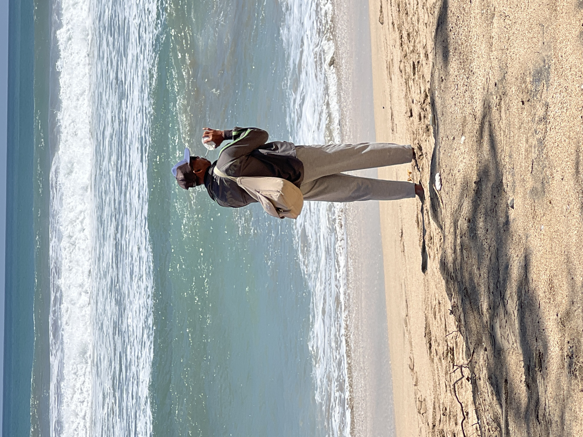 A man stands on a sandy beach in Kuta, Indonesia, facing the ocean. He is wearing a grey jacket with green accents, light grey pants, and a blue baseball cap. A light brown shoulder bag is slung across his back. He is holding a white cup to his mouth, appearing to drink from it. The ocean in front of him is a vibrant turquoise with white-capped waves. The sky above is a clear blue. The beach is composed of light brown sand with some scattered dry branches and shadows cast by unseen objects. A white piece of trash is visible on the sand near the man's feet.