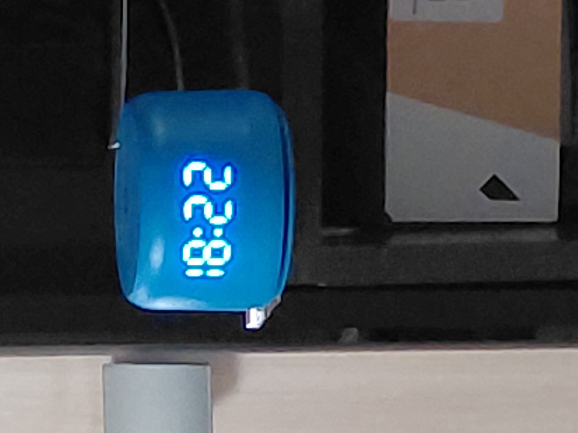 A small, blue, rectangular digital device with rounded edges is prominently displayed in the center of the image. Its bright blue LED screen shows the digits "22:88" oriented vertically, with "22" at the top and "88" at the bottom, separated by a colon. A thin, light-colored wire is visible, connecting to the upper-left side of the device. The device appears to be resting on a light-colored, flat surface, possibly a shelf or table. To the right, a rectangular object resembling a cardboard box or part of a piece of furniture is partially visible. This object has a diagonal design, with its upper section being light brown or orange and its lower section a lighter gray or white, featuring a small black triangular cutout. Some faint, unreadable text is present on the light brown section of this object. The background is dark and out of focus, suggesting an indoor setting. No discernible activity or event is taking place. The time of day or specific function of the "22:88" display is not clear, as it doesn't conform to a standard clock format. The location context is Nadym, Russia.