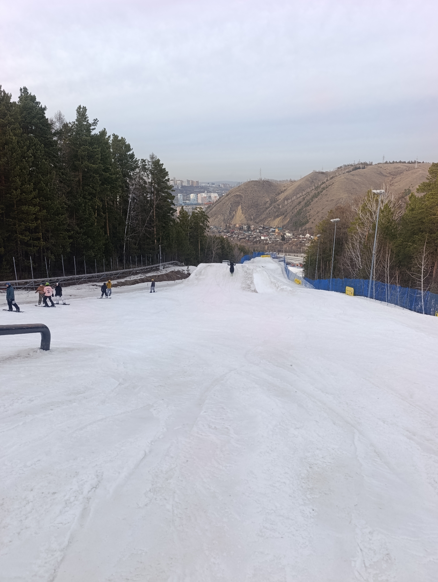 The image shows a snow-covered slope, likely for skiing or snowboarding, at a ski resort. In the background, there is a landscape with trees, a city skyline, and hills. Several people are visible on the slope, some standing and others in motion, presumably skiing or snowboarding. A blue safety fence runs along the right side of the slope. There is a small jump constructed on the slope. The environment is cold and overcast. Based on the landscape and the presence of the ski resort, the location is likely within or near Krasnoyarsk, Russia, as suggested by the query.