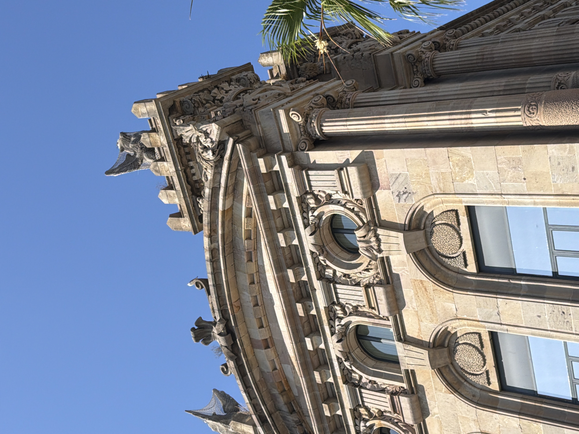 The photograph captures an upward view of an ornate building facade against a clear blue sky. The building is constructed of stone and features classical architectural elements such as columns, arches, intricate carvings, and statues. The top of the building is adorned with battlements and a sculpted figure that resembles a winged creature. A small bird is perched on the roofline. The lower portion of the building displays arched windows with decorative surrounds. A palm frond is visible in the upper right corner of the image, suggesting a warm climate. The architecture is reminiscent of modernist or art nouveau styles, commonly found in Barcelona.