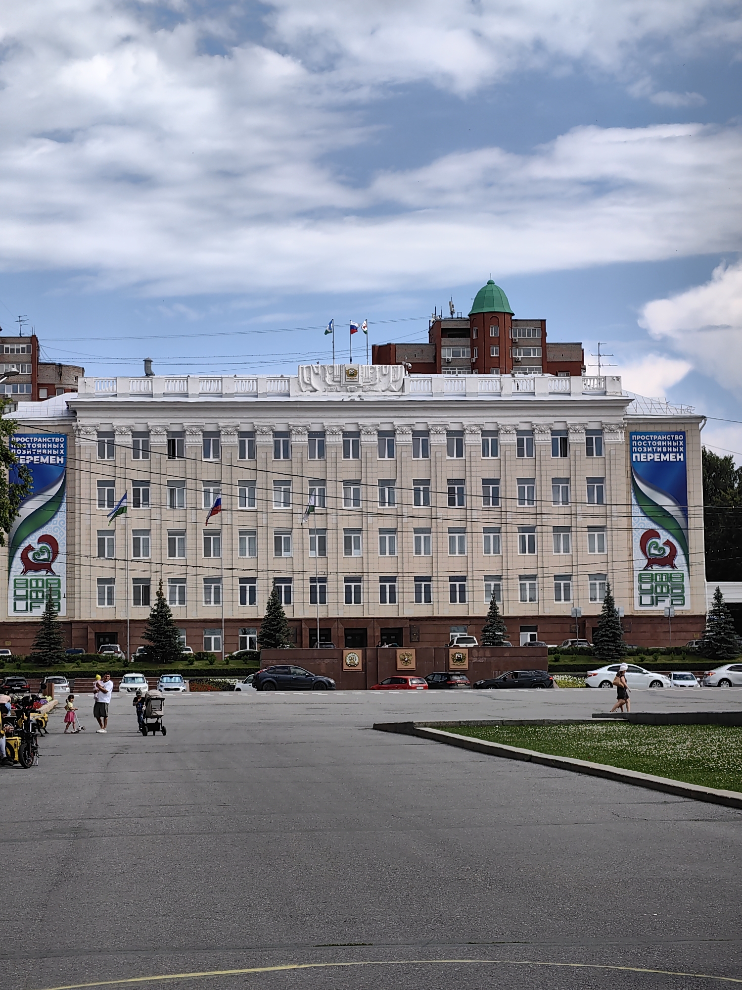 A wide shot captures a grand, light-colored building with many windows, suggesting a civic or governmental structure, in Ufa, Russia. The building is topped with a white balustrade and features prominent columns along its facade. Large banners with blue and green designs hang on either side of the central section, displaying Cyrillic text. The banner on the left reads "ПРОСТРАНСТВО ПОСТОЯННЫХ ПОЗИТИВНЫХ ПЕРЕМЕН" and the one on the right has similar text.
In front of the building, a paved plaza is visible, with several cars parked and driving. A few people are present on the plaza, including a family with children and a woman walking alone. Some trees are planted in front of the building and along the edge of the plaza. In the background, another building with a distinctive green dome is visible. The sky is partly cloudy, indicating daytime. The overall scene conveys a sense of public space and civic presence.