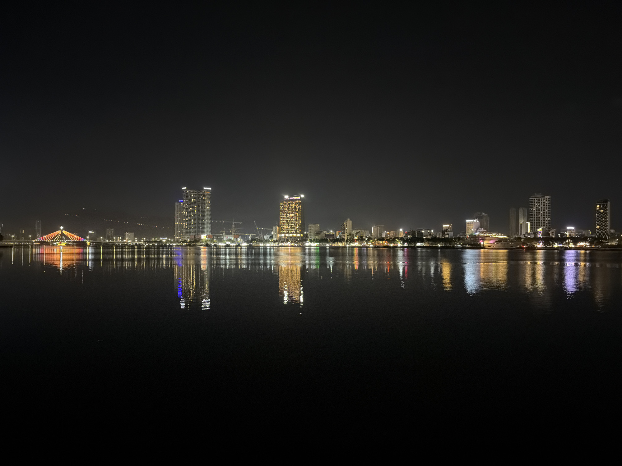 The image depicts the skyline of Đà Nẵng, Vietnam, at night, reflected in the calm surface of the Han River. The scene is dominated by various lit buildings and the Dragon Bridge (Cầu Rồng), recognizable by its distinctive arch and colorful illumination. The water surface shows distinct reflections of the buildings and bridge lights. The sky is dark and free of visible clouds. The foreground is largely dark, emphasizing the skyline's illuminated elements and their watery reflection.