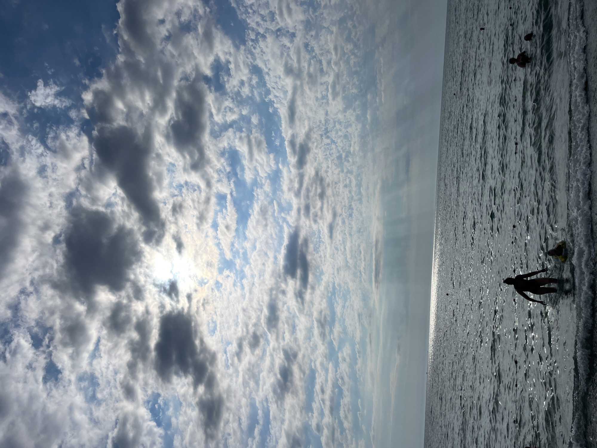 A silhouetted figure stands in the shallow water of the sea, facing away from the camera, with another figure in a yellow float nearby. The scene takes place in Sochi, Russia, on a cloudy day. The sun is partially obscured by clouds, casting rays of light onto the water. The sea is choppy with small waves breaking on the shore. In the distance, other people are visible swimming in the sea. The overall atmosphere is one of leisure and relaxation.