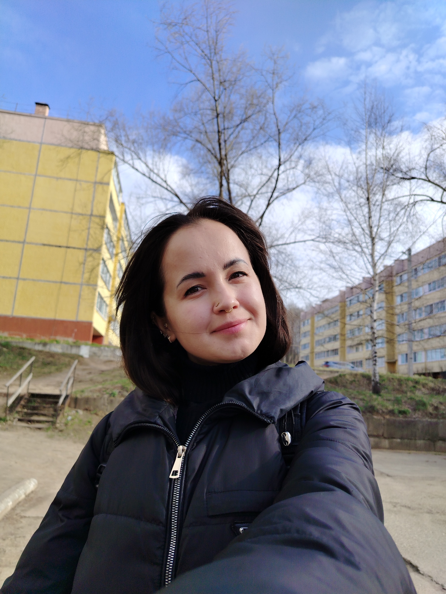 A medium shot depicts a woman with fair skin and dark brown hair, dressed in a black puffer jacket and black turtleneck, smiling slightly at the camera. She has a nose piercing on the left side. In the background are two yellow buildings with multi-paned windows, and bare trees against a blue sky with scattered clouds. A set of wooden stairs is visible to the left. The scene appears to be outdoors, possibly in a residential area. Based on the provided information, the location is Noviy, Russia.