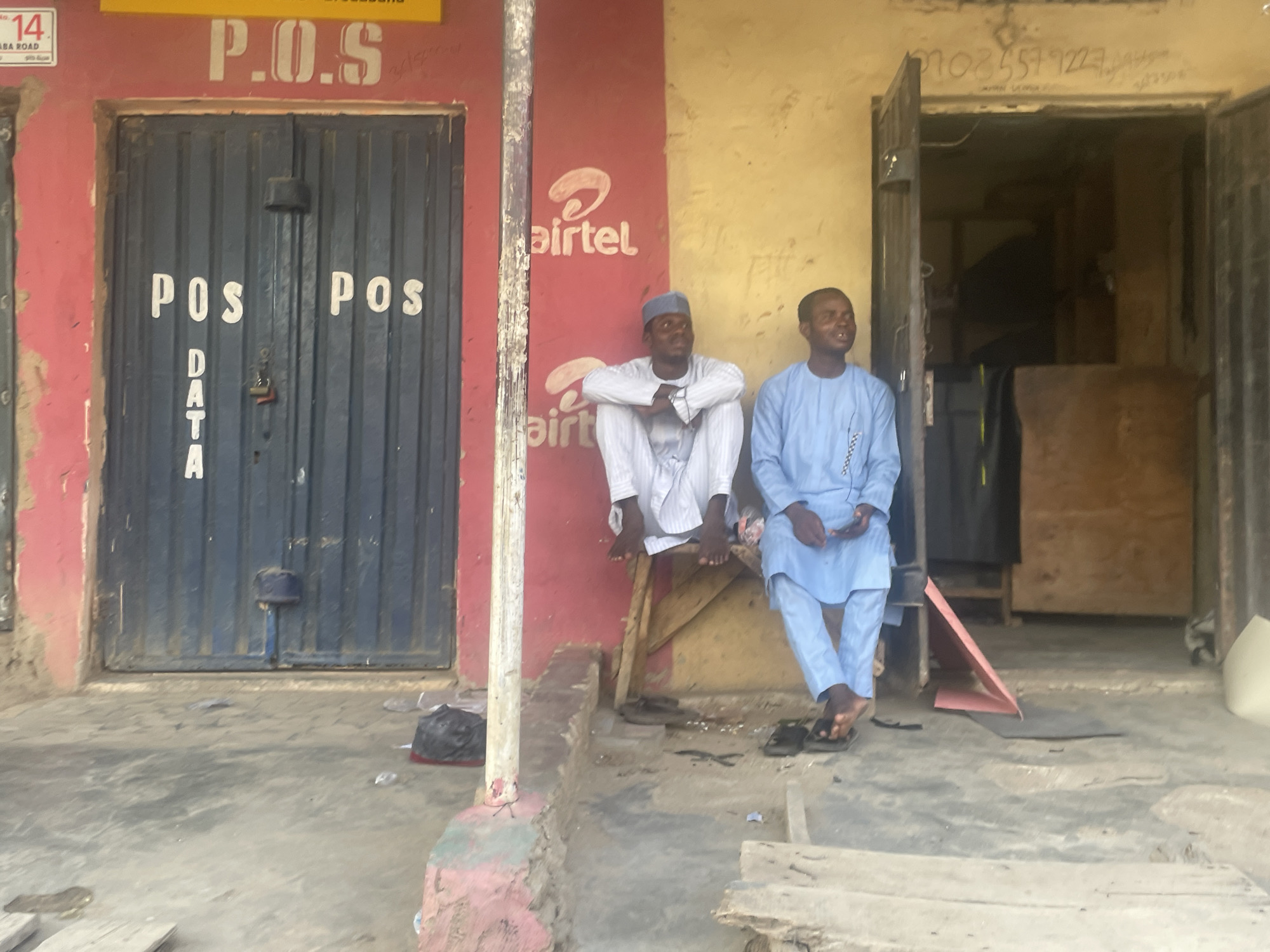 Two men are seated outdoors on a concrete pavement, in front of a building with two adjacent storefronts. The scene appears to be at street level during bright daylight.
On the left, a vibrant red wall features a dark blue corrugated metal door. Painted on the door in white are the words "POS DATA," with "POS" repeated on each half of the double door. Above the door, a yellow sign with red lettering displays "P.O.S." Another small white sign to the upper left of the door reads "No. 14 SABA ROAD." Further right on the red wall, the white "Airtel" logo is prominently painted. A weathered white-painted pillar stands between the red and a yellowish wall.
The man on the left is dressed in a white traditional outfit and a matching skullcap, sitting on a low wooden stool, with his arms crossed and hands tucked into his sleeves. He looks slightly towards the right with a faint smile. His bare feet rest on the stool. The man on the right, clad in a light blue traditional tunic and trousers, sits on a similar low wooden structure, looking upwards and to the right with a broad smile. His black flip-flops are on the ground near his bare feet.
To the right of the men, an open doorway reveals a dimly lit interior. Above this doorway, some faded handwritten numbers, including "0805571223," are visible. The walls show signs of wear and tear with peeling paint. Scattered on the concrete ground are a few items, including what appears to be a black plastic bag and some debris. The overall impression is that of a bustling, lived-in commercial area.