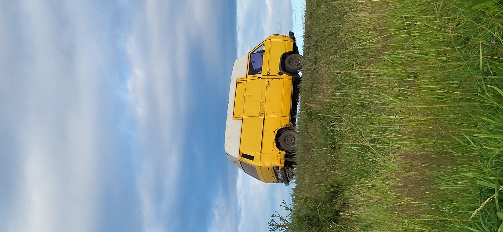 A yellow and white van is parked on a grassy hill under a cloudy sky. The van is oriented perpendicular to the viewer, with its front facing right. The van is weathered and appears to be an older model. The grass in the foreground is tall and green, with some dry stalks mixed in. The sky is filled with large, grey and white clouds, with patches of blue visible. The scene appears to be in a rural or undeveloped area, possibly a field or roadside in Hetmanivka, Ukraine. There are no people visible in the image.