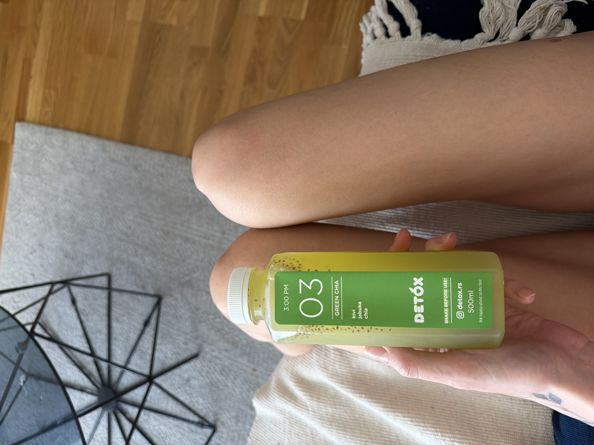 A woman is seated with her thighs together, holding a bottle of green detox juice. The bottle is labeled "3:00 PM 03 GREEN CHIA kivi jabuka chia DETOX SHAKE BEFORE USE! detox.rs 500ml Rok trajanja utisnut na dnu boce." The juice appears to contain chia seeds. The setting includes a wooden floor, a light grey rug, and a patterned cushion or blanket. There is a decorative geometric metal object on the rug. Based on the brand "Detox.rs", the location could be in the city of Belgrade, Serbia.