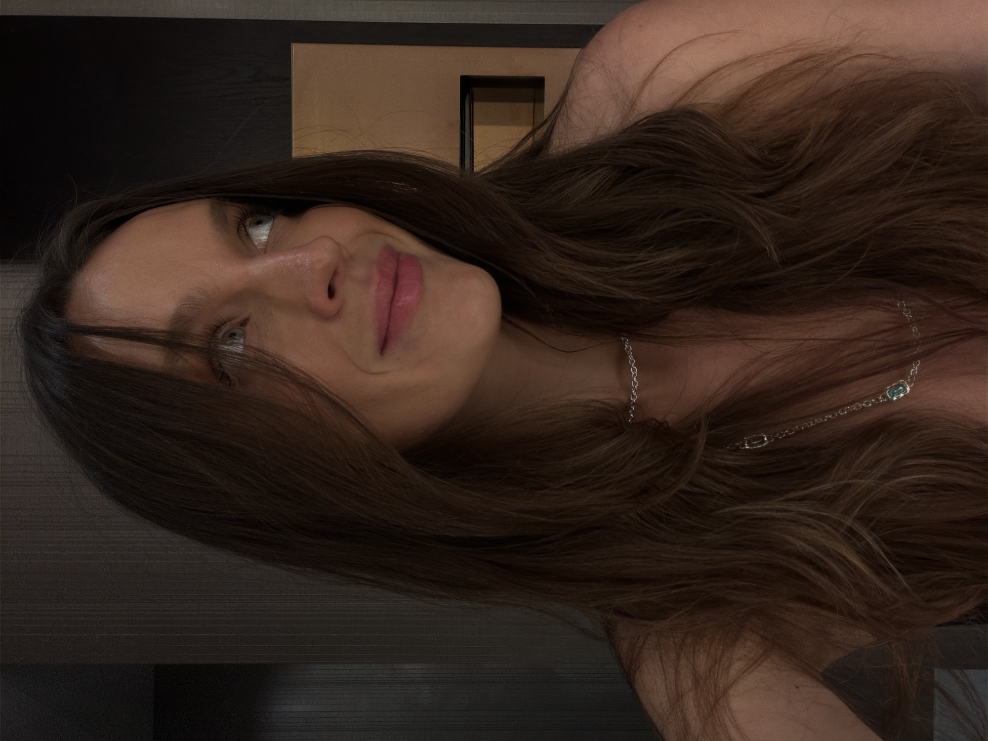 A woman with long, light brown hair and light-colored eyes is positioned at a low angle, giving the impression that she is lying down or looking upward. She is wearing a silver necklace with what appears to be a blue stone, and is positioned indoors against a dark surface that suggests a table or another piece of furniture. In the background, a black and tan colored door is visible. The location in the city of Budva, Montenegro cannot be determined from the image.