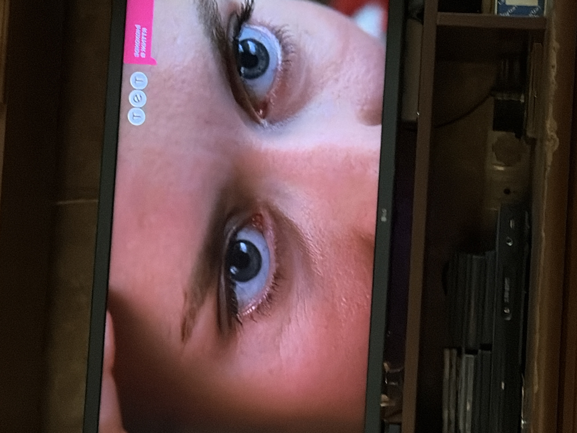 The image shows a close-up view of a person's eyes, with a television screen in the foreground. The person's eyes are open and looking forward, with visible eyelashes and eyebrows. The skin around the eyes appears slightly flushed. On the top right corner of the TV screen, there is a logo with three circles and a pink rectangular banner with text in what appears to be Cyrillic script. Below the television, there is a shelf with stacked items, possibly DVDs or game cases, and what looks like a DVD player or similar electronic device. The overall lighting suggests the image was taken in an indoor setting, likely a living room or media room. There is no discernible urban or geographical information present in the image to indicate a specific city or location.