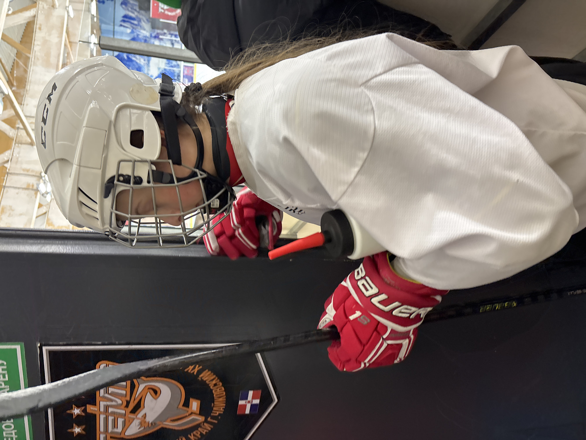 A person wearing a white CCM hockey helmet, red Bauer gloves, and a white jersey holds a hockey stick. The stick is resting on a black surface, possibly a bench or dasher board. A black rectangular sign with a logo featuring a stylized bird and stars is visible, along with text in Cyrillic script. A small flag of the Dominican Republic is visible on the sign. The location is in Chaikovsky, Russia, according to the text on the sign. The person appears to be preparing to play hockey.