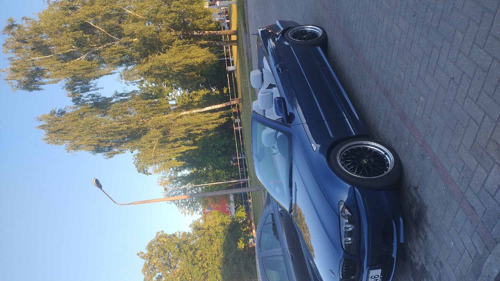 In the city of Jelgava, Latvia, a blue BMW convertible is parked in an outdoor lot. The car has a light-colored interior and black rims. Next to it, a dark-colored sedan is also parked. The background features tall trees with green foliage and a bright blue sky, indicative of a sunny day. A lamppost is visible on the left side of the image. The ground is paved with interlocking brick pavers. A red line is marked on the pavers near the car.