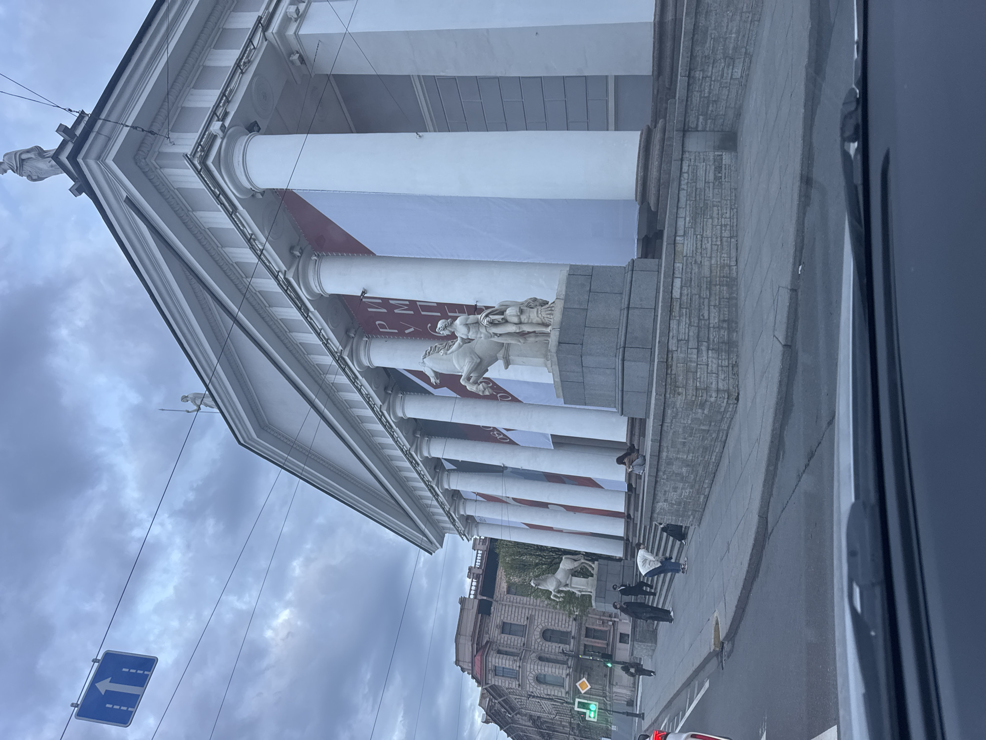 The image shows a view of the Mining University in Saint Petersburg, Russia. The building has a neoclassical facade with tall white columns and statues on the roof and above the entrance. A large white banner with red accents is hung between the columns. Pedestrians walk on the sidewalk in front of the building. Traffic lights show green. A blue road sign is visible in the sky. The sky is overcast. The view is partially obstructed by the interior of a car.