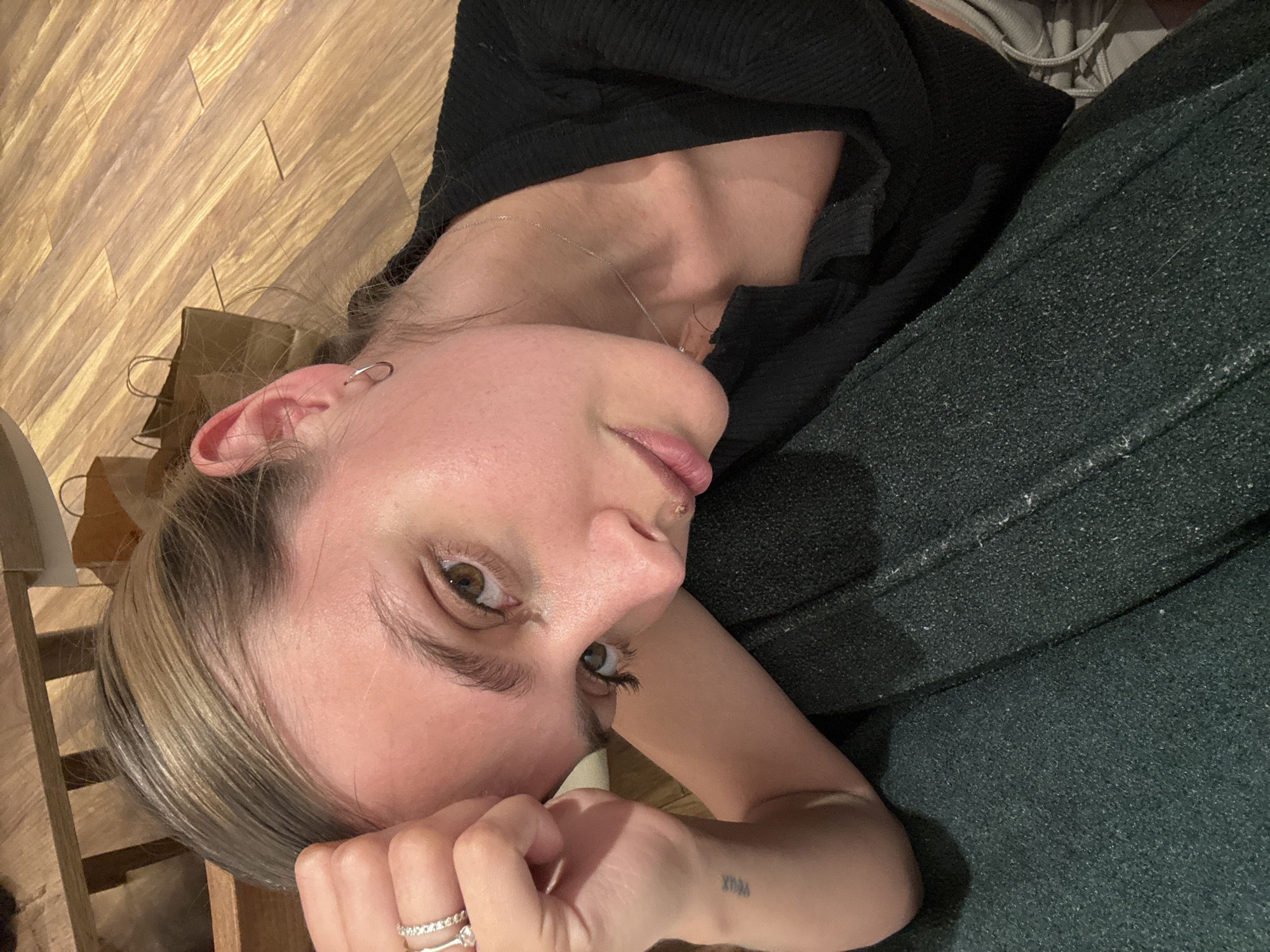 A person lies on a wooden floor, their head close to a wooden structure. They are wearing a black top and green pants or a blanket. A small tattoo is visible on their arm. A ring is visible on their finger. The location is Unknown, Unknown.