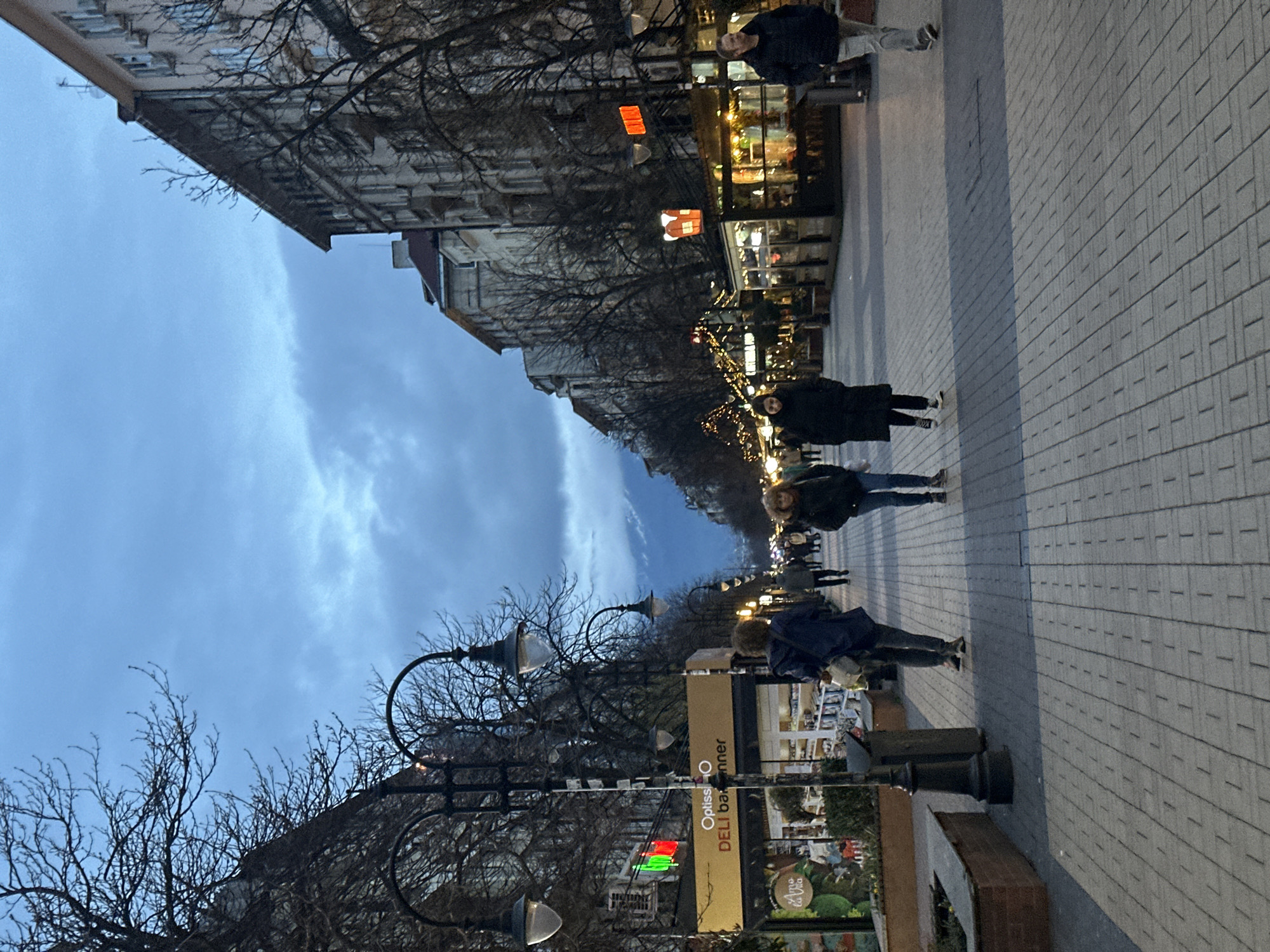 The image shows a street scene in Sofia, Bulgaria. The street is paved with grey rectangular stones. There are several people walking along the sidewalk, including one person near the top right corner of the image and two figures further down the walkway. A large building is visible on the left side of the street, and shops with illuminated windows are seen on the right. Bare trees line the street, with decorative lights strung among them. There are vintage street lamps and advertisements along the street. The sky is overcast. There is a sign for "Optissio DELI banner".