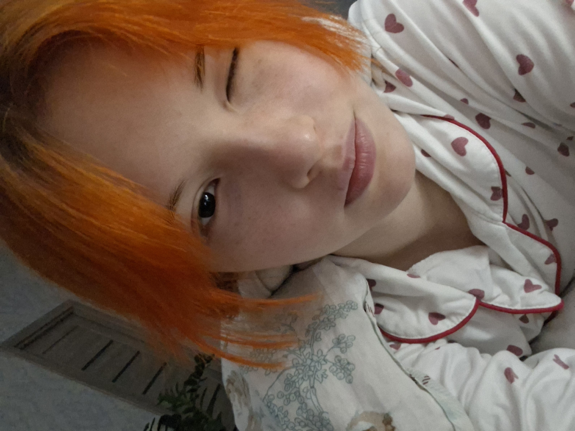 A young person with short, vibrant orange hair winks at the camera. The person's skin is light, with visible freckles. They are wearing a white pajama top with small pink hearts and red piping. The pajama top is covering what appears to be a pillow with a light-colored pattern. The background is slightly out of focus, showing a vent on a wall, a plant, and a door. There is no information available to confirm the location as Chaikovsky, Russia.