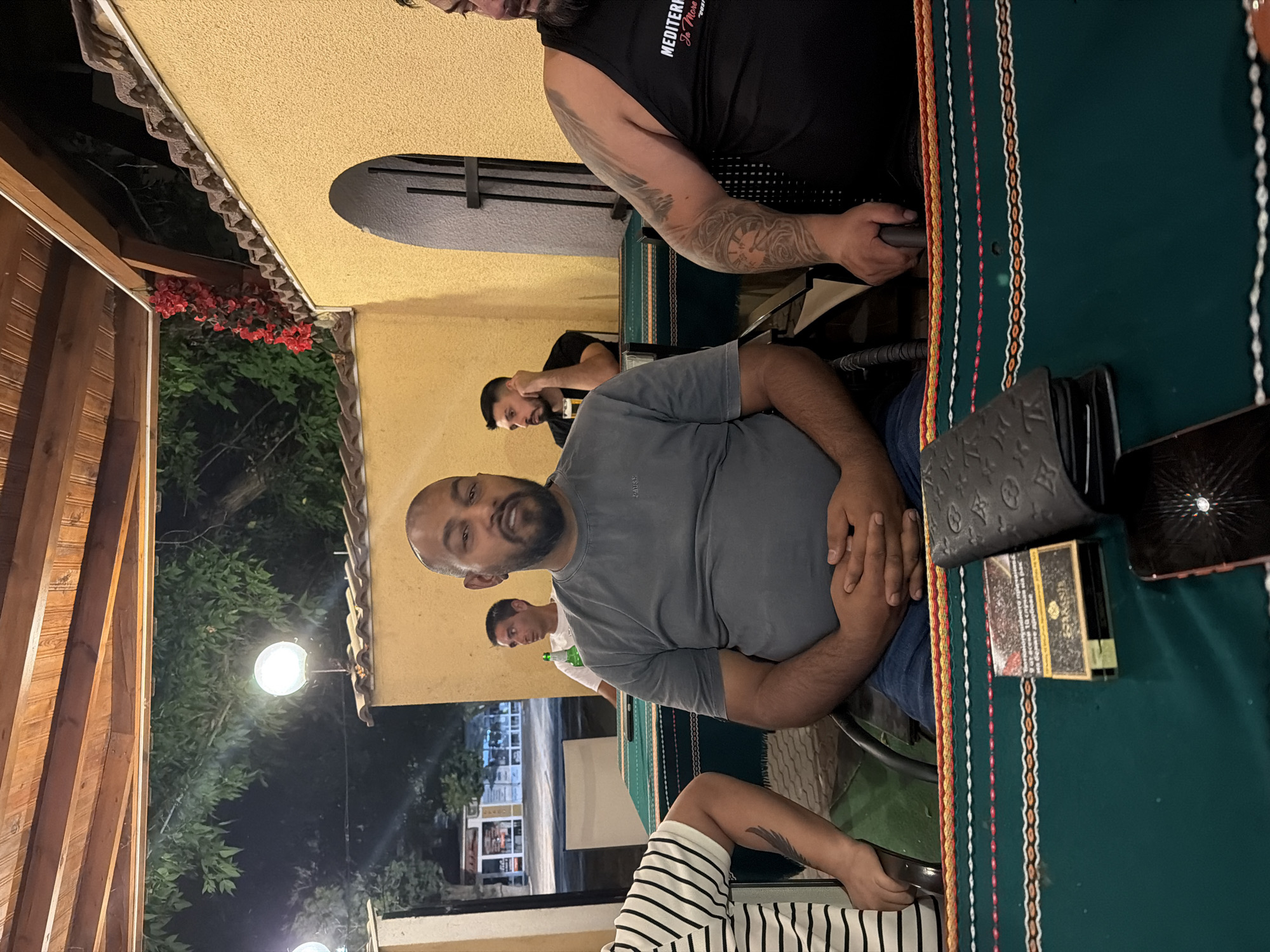 The image depicts a group of men at an outdoor dining establishment in Levski, Bulgaria. The central figure, a bald man with a beard, is seated at a table and looking directly at the camera. He is wearing a gray t-shirt. To his left, another man with a striped shirt is partially visible. To the right of the central figure, a man in a black tank top with visible tattoos is seated. In the background, two other men are visible, one holding a glass and the other looking away.
The table is covered with a green tablecloth featuring colorful embroidered patterns. On the table, a pack of cigarettes and a wallet are visible. The setting appears to be a restaurant or cafe with wooden roofing and yellow walls. Red flowers are visible in the background. The overall lighting suggests it is evening or nighttime.