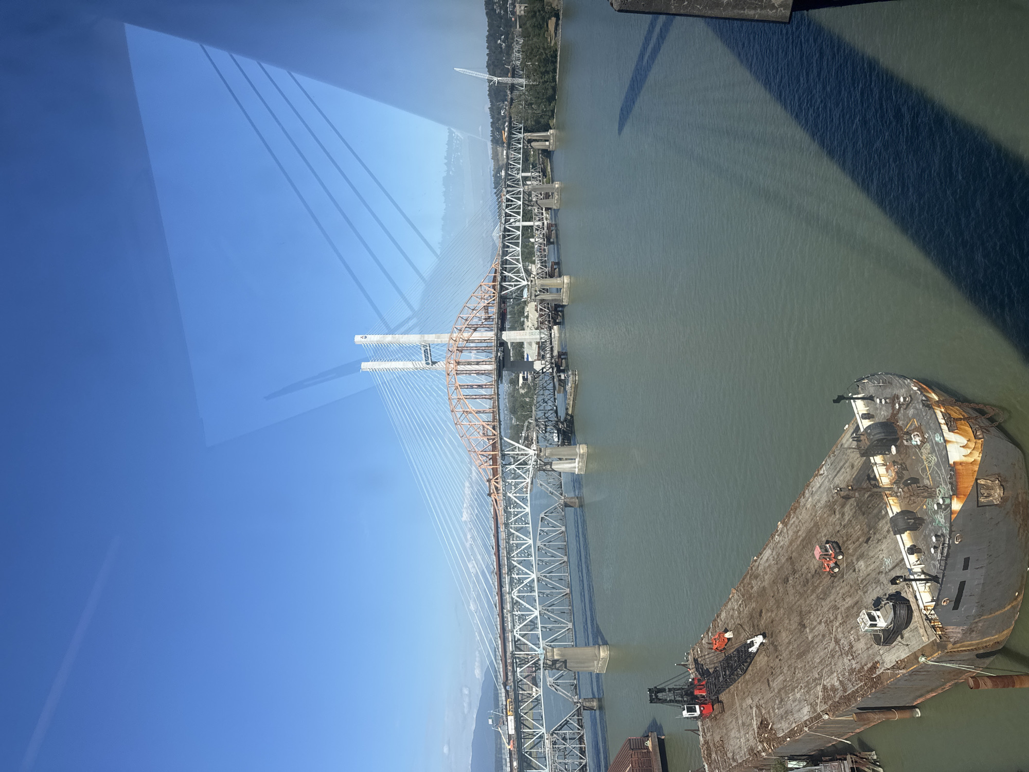 This image shows a wide, greenish-blue body of water, likely the Fraser River, with several bridges crossing it. In the foreground, a large, flat-topped barge is moored. The barge is weathered and rusty, with several pieces of construction equipment on its deck, including a red and white crane, a smaller orange vehicle, and a white tugboat. The barge is positioned to the left of the frame.
Dominating the mid-ground are two bridges. The closest bridge is a multi-span truss bridge with concrete piers. Above this, a more modern cable-stayed bridge is visible. This bridge has tall white towers and an orange-colored arched section. Vehicles are visible on the upper bridge. In the background, the shorelines of New Westminster are lined with trees and buildings. Distant mountains can be seen on the left side of the frame. The sky is clear and bright blue, with reflections from the sky and a structure visible on the surface of the water. The overall scene suggests an industrial waterfront area in New Westminster, Canada, with ongoing construction or port activity.