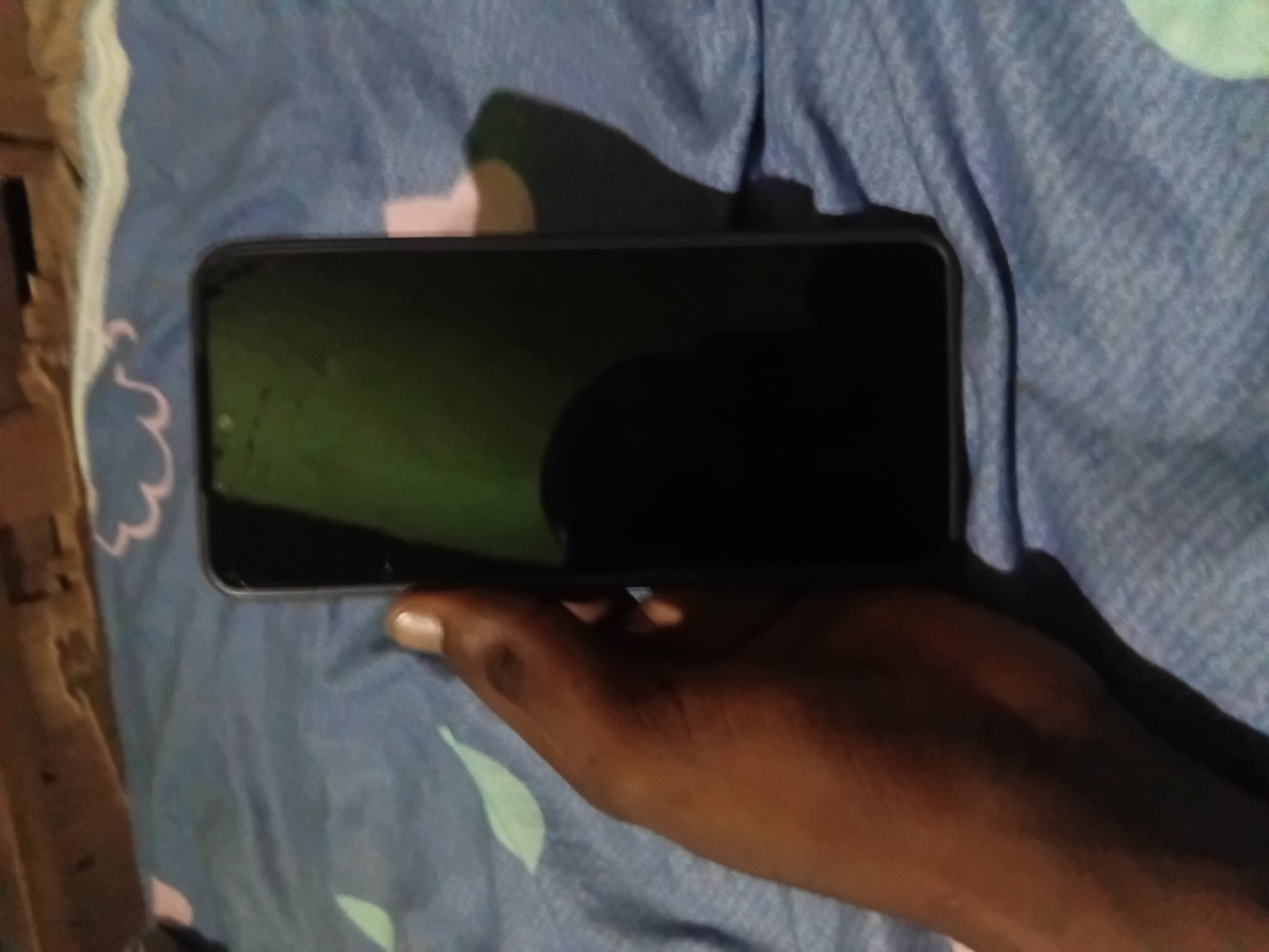 A person's dark-skinned hand is holding a black smartphone with a dark screen. The phone is held vertically, angled slightly towards the viewer. The background is a blue fabric with a subtle pattern of white and light green shapes, resembling stylized leaves or clouds. Above the fabric, a wooden surface with a dark, textured finish is visible, possibly a table or shelf. The lighting in the image is dim, casting a shadow of the hand and phone onto the blue fabric. The scene appears to be an indoor setting.