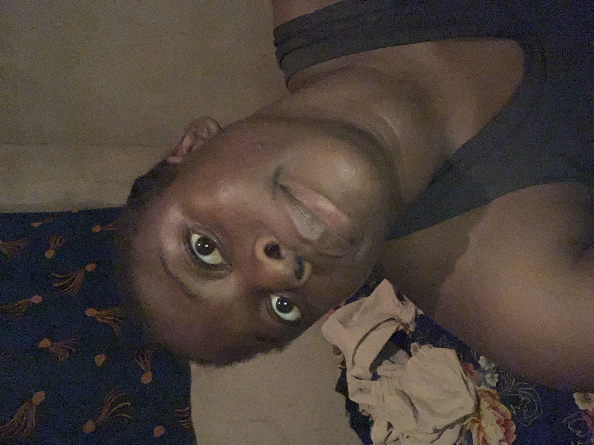 A close-up, indoor shot shows a dark-skinned person with a shiny forehead and visible pores, lying on what appears to be a patterned bed covering. The person's face is turned towards the camera. Clothing is visible on their shoulder and chest. A pile of light-colored fabric is near their torso. The environment is dimly lit. The location in the city of Agban, Nigeria cannot be determined from the visual information provided.