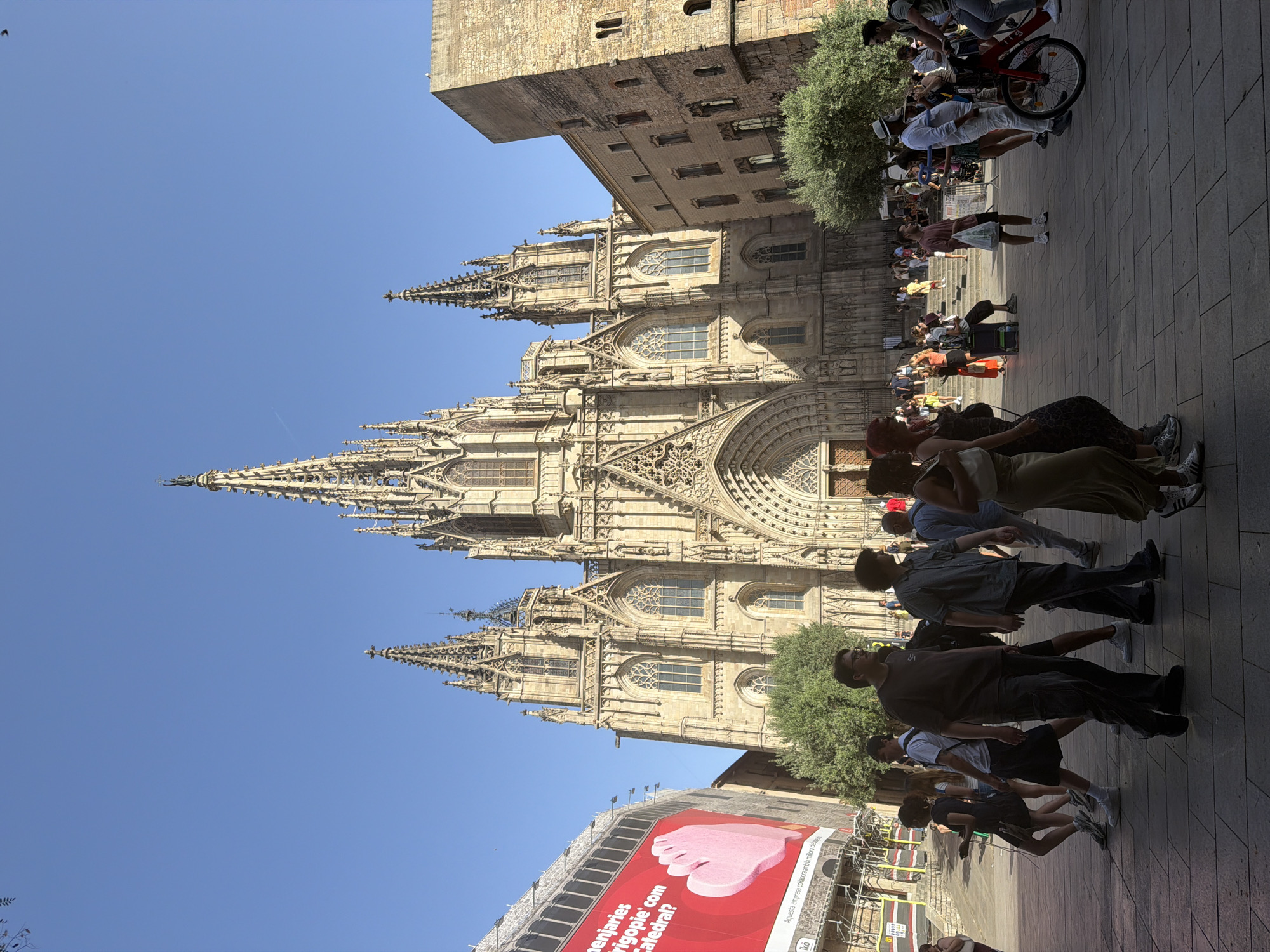 The image captures a street scene in Barcelona, Spain, with the Barcelona Cathedral as the dominant backdrop. The cathedral, a grand Gothic structure, features intricate stonework, tall spires, and large arched doorways. In the foreground, a paved plaza is populated with a diverse group of people, some walking, others standing and observing. A large red banner with white text and a pink graphic is visible on the left side of the image, partially obscuring a modern building. The sky is clear and blue.
Among the people, several individuals are in focus: a man wearing a dark t-shirt and cargo pants is walking towards the right, a man in a blue checkered shirt and dark pants is walking in the same direction, and a woman in a long, flowing dress is also moving to the right. Further in the background, more individuals are visible, some appearing to be tourists, while others might be locals. A red bicycle with a white basket is parked on the right side of the frame. The lighting suggests it is daytime, with shadows cast by the buildings and the people.