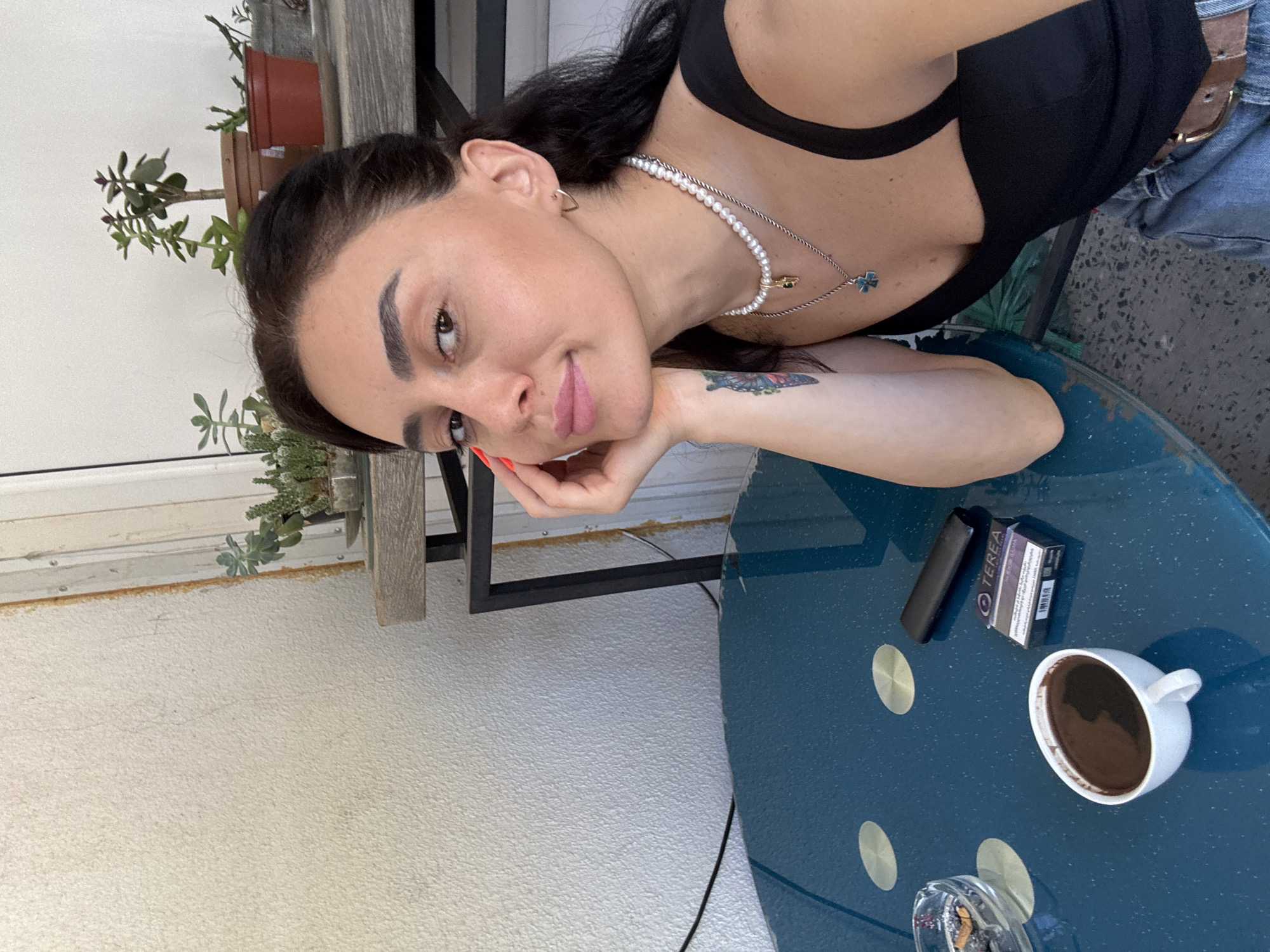 A woman is taking a selfie at a table outdoors, with a view of several potted plants on a shelf behind her. She has dark hair and is wearing a black tank top, a pearl necklace, and jeans. On the blue table in front of her are a white cup of coffee, an ashtray with cigarette butts, a pack of TEREA cigarettes, and a black device, possibly an e-cigarette. The environment appears to be a cafe or restaurant in Tbilisi, Georgia, given the presence of plants often found on balconies or patios in the city, and the distinct stucco texture of the building's exterior wall, a common architectural feature in Tbilisi.