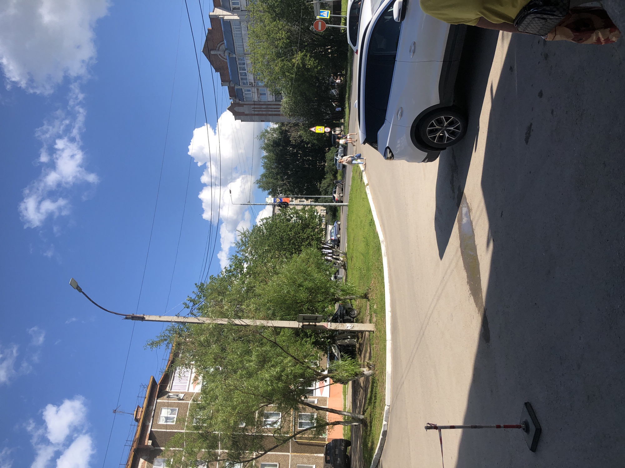 The image depicts an outdoor scene on a sunny day with a bright blue sky and scattered white clouds. The perspective is from a slightly elevated viewpoint, looking down towards a street and sidewalk. A white car is parked on the right side of the frame, with its right rear wheel visible. The car's shadow stretches across the asphalt.
In the background, there are apartment buildings, some with visible windows and balconies, and a mix of mature trees and greenery. A street lamp with a curved arm is positioned on the left side of the image, casting a shadow. Power lines are strung across the sky.
Further down the street, several people are walking. Two individuals, appearing to be women, are seen walking on the sidewalk near a yellow traffic sign with a pedestrian symbol. Another person, possibly a woman, is walking closer to the foreground, carrying a bag. A red and white striped barrier pole with a square base stands on the asphalt in the lower right corner of the image.
The overall scene suggests a typical urban environment in Solikamsk, Russia, during daylight hours. The weather appears to be warm and pleasant, indicated by the clear sky and shadows. The presence of people and vehicles suggests normal daily activity. There is no visible text in the image that can be clearly discerned or interpreted for meaning.