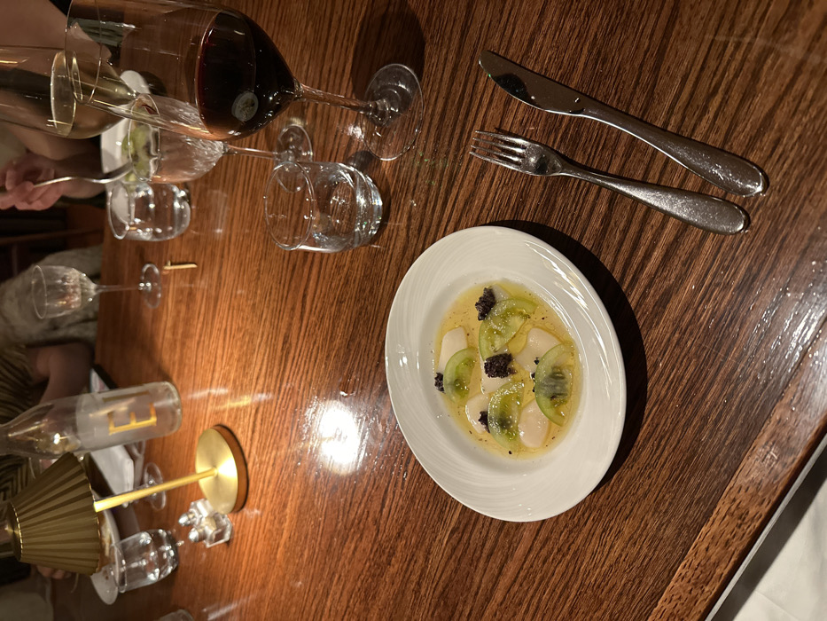 The image depicts a dining table scene at The Wolseley restaurant, located at 160 Piccadilly, St. James's, London W1J 9EB, United Kingdom. The primary surface is a dark, polished wooden table exhibiting a distinct grain pattern, illuminated by warm, localized lighting. In the foreground, a white oval ceramic plate holds a dish composed of thinly sliced pale, translucent items, possibly scallops or radish, arranged in a shallow pool of light yellow liquid. These are garnished with multiple thin slices of green fruit, consistent with kiwi or unripe tomato, and several small, dark, granular clusters resembling olive tapenade or caviar. To the right of the plate, a silver dinner fork and knife are placed parallel to each other. Further right, an empty small glass tumbler and a tall stemmed glass containing red wine are visible. In the midground, additional glassware includes a stemmed glass with white wine and another water glass. On the left side of the table, a clear glass bottle with a prominent "E" logo is partially filled with water, positioned next to a small, gold-colored, pleated table lamp emitting a warm glow. Two small, clear salt and pepper shakers are located behind the lamp. Blurred figures of individuals are discernible in the background, one with a hand holding a fork, indicating active dining.