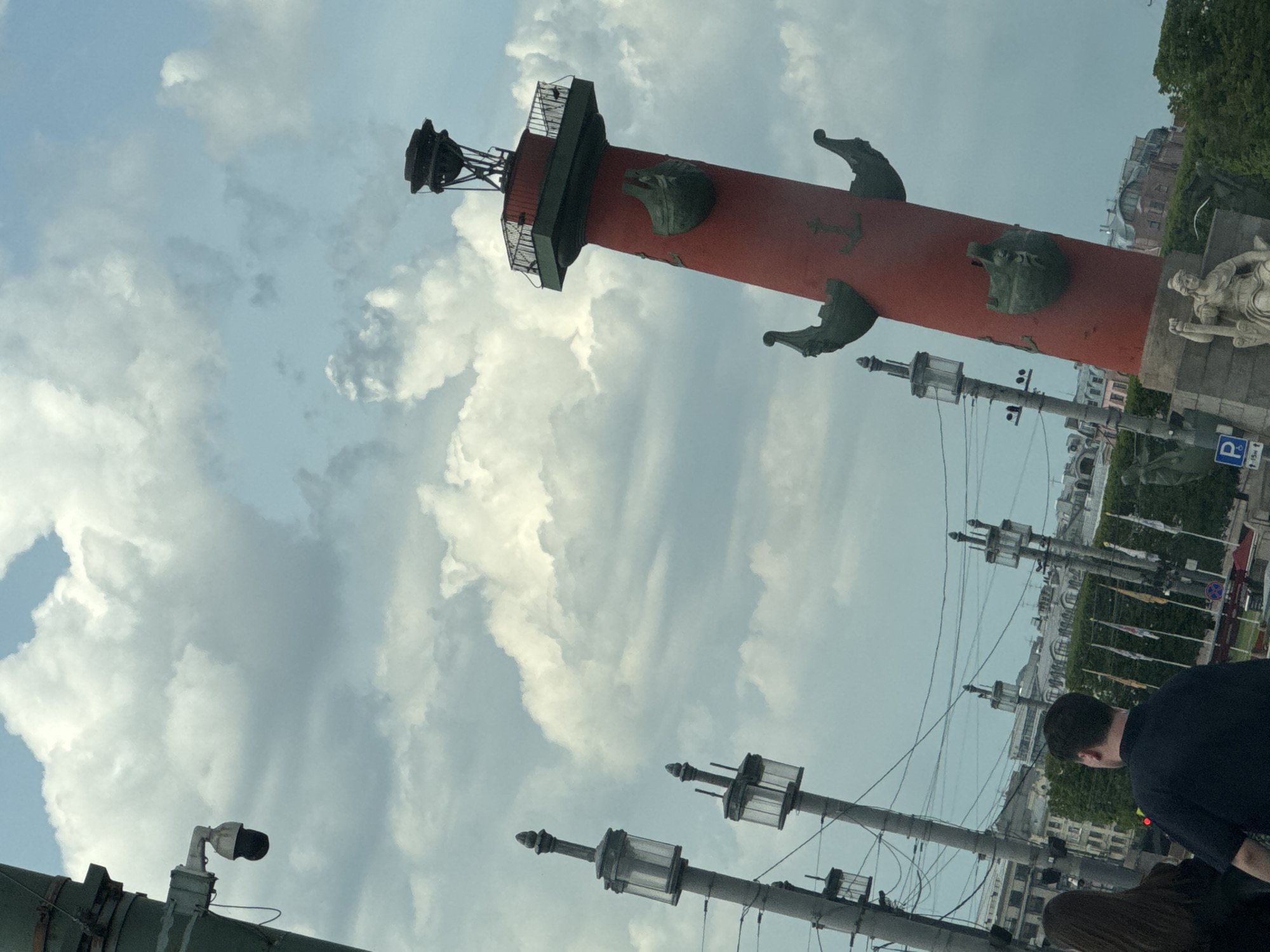 The image displays a tall, red Rostral Column, prominently featured against a cloudy, bright sky in Saint Petersburg, Russia. The column, a distinctive landmark often found on the Spit of Vasilyevsky Island, is adorned with bronze ship prows (rostra) and anchors along its length. Near its top, there is a dark observation platform or balcony, capped by a metal structure that historically functioned as a beacon or censer.
In the foreground and midground, multiple street poles, streetlights, and a web of overhead wires are visible. To the right of the column, a large, light-colored stone statue of a draped, seated figure on a pedestal is partially captured, representing one of the allegorical figures often associated with these columns. The back of a person's head and shoulders, wearing a dark shirt, is seen on the far right, with a glimpse of another person's hair below, indicating human presence in the urban setting.
The background reveals classical-style buildings with multiple stories and arched windows, typical of Saint Petersburg's architecture, along with some green foliage from trees. The sky is largely covered with large, white and grey clouds, suggesting a cloudy but well-lit day. A blue parking sign with a white "P" symbol is visible on the right side of the frame. The scene captures a historical monument within an active urban environment, likely in the daytime.